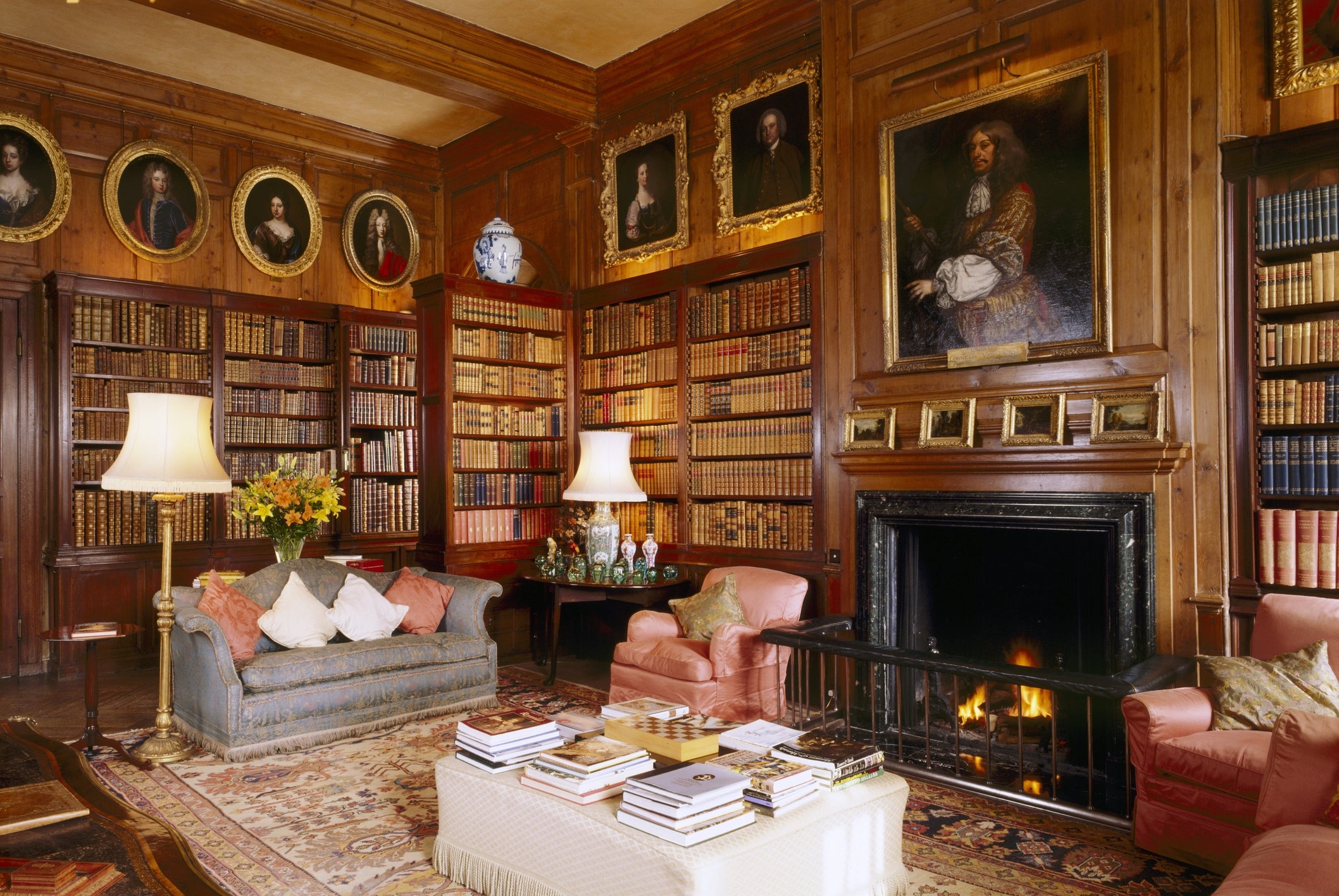 Luxurious home library with wooden shelves filled with books, antique portraits, a fireplace, and elegant seating around a coffee table
