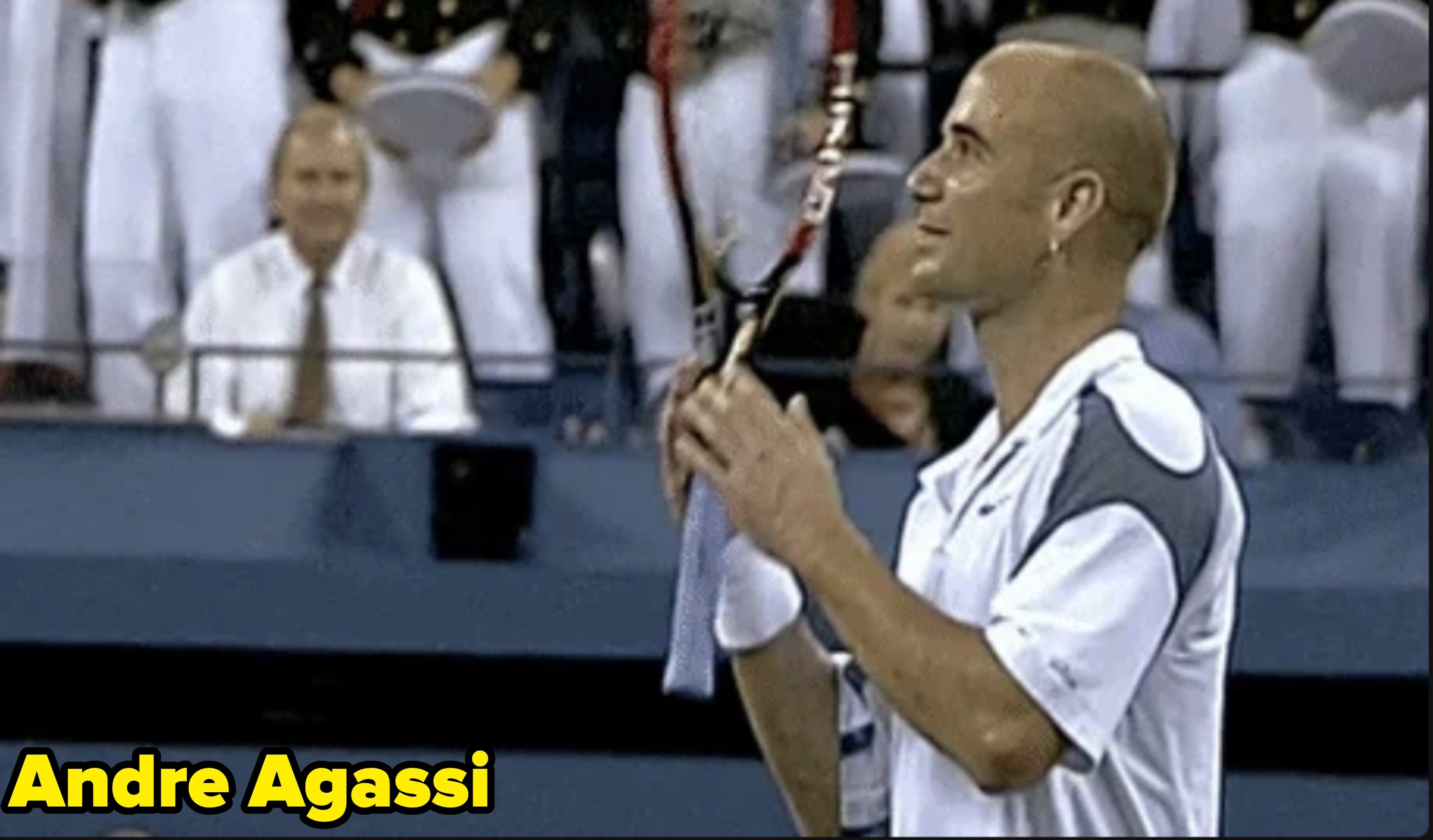 Athlete with a shaved head, wearing a tennis outfit, holding a racket on a tennis court, surrounded by seated spectators