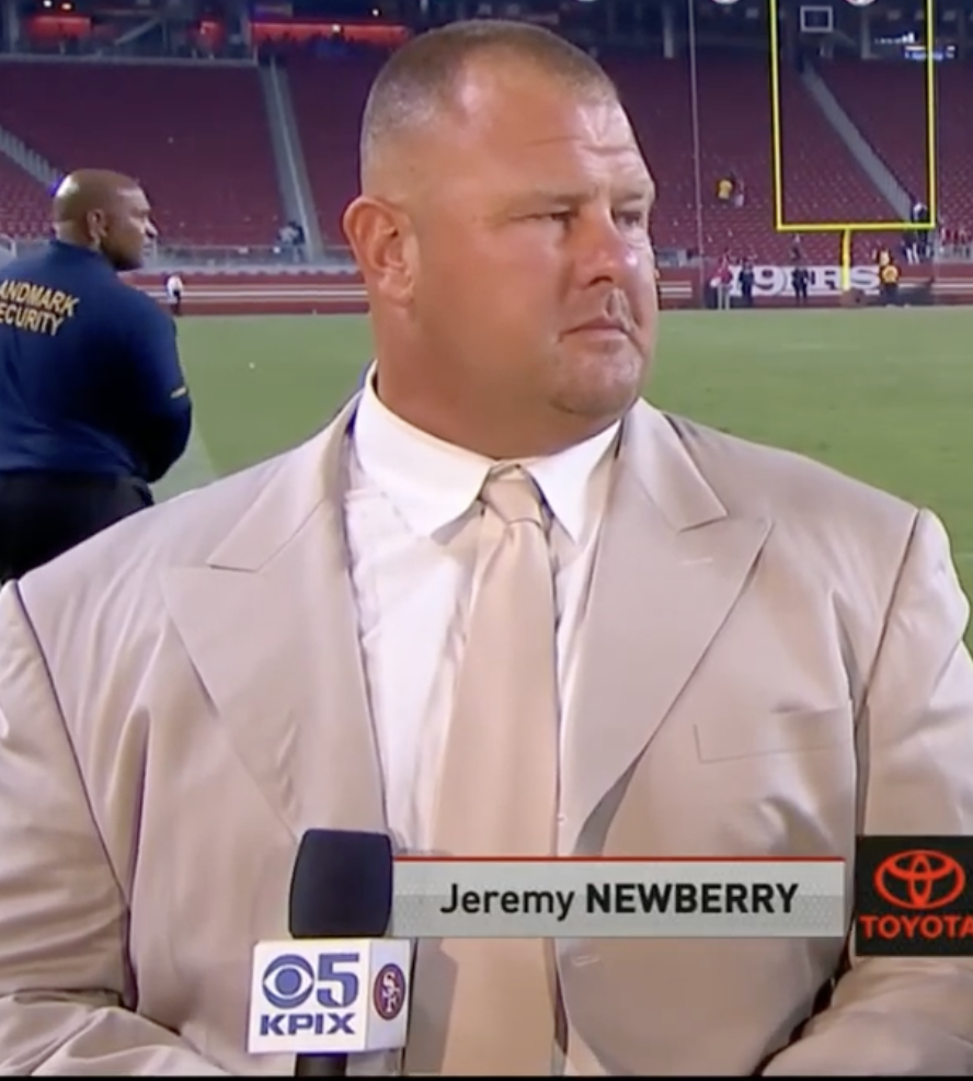 A person in a suit stands on a football field, being interviewed for TV