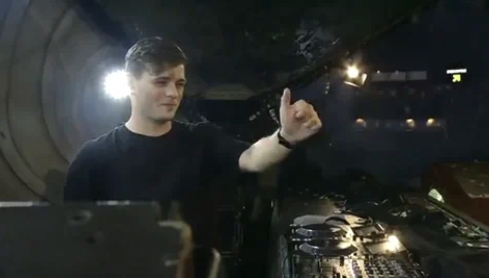 A DJ performing live, giving a thumbs-up gesture while standing at a deck with equipment in a dimly lit venue