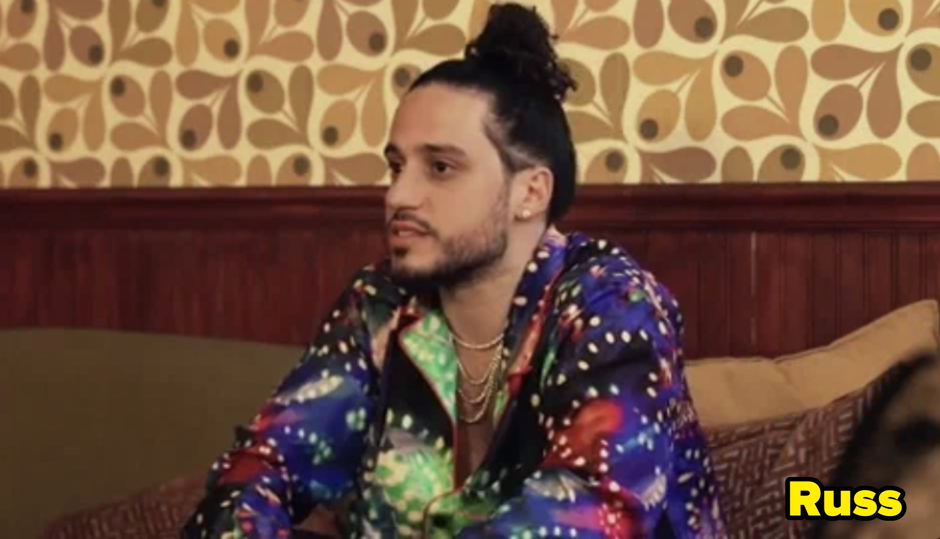 A person in a patterned jacket sits on a couch, looking to the side, with a wallpapered background
