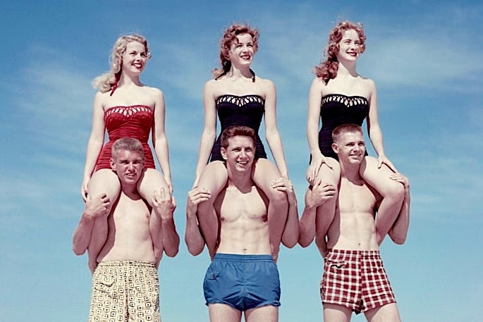 Young adults in 1950s swimwear pose on a beach, each woman on a man's shoulders, surrounded by sand and grass under a clear sky