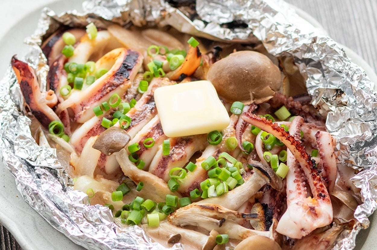 ホイルに包まれたイカとキノコのバター焼きが皿に盛られている。青ねぎがトッピングされている。