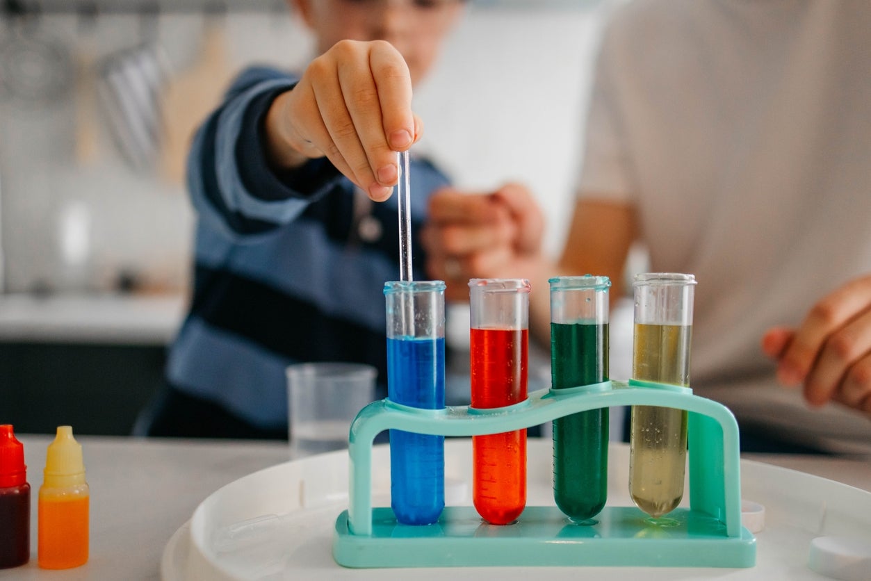 Child holding a dropper above test tubes with different liquids, engaging in a science activity. Adult hands are nearby offering guidance
