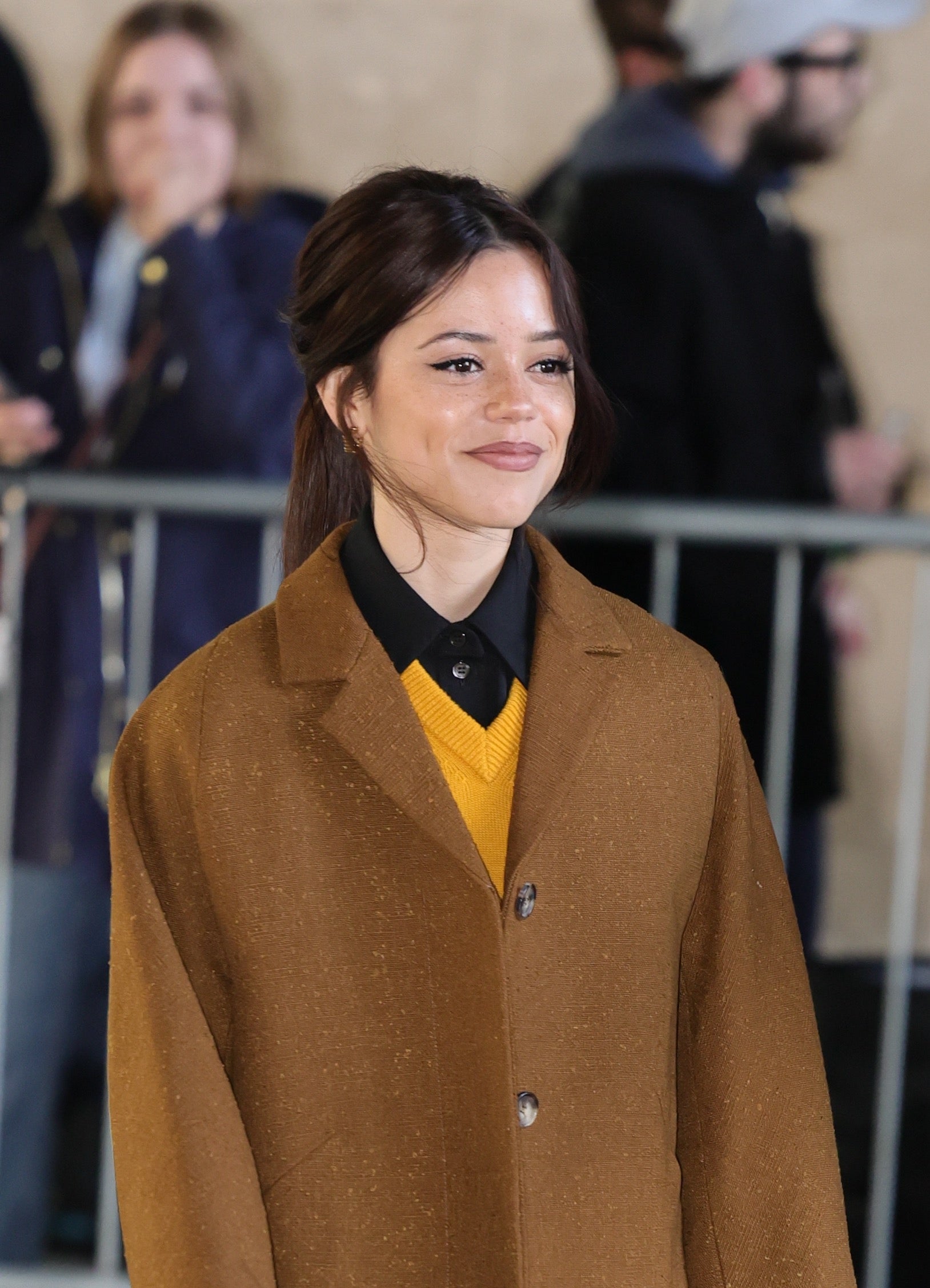 Jenna Ortega’s natural-looking brow definition Close view of Jenna Ortega smiling with darker brows and a brown coat at a London appearance