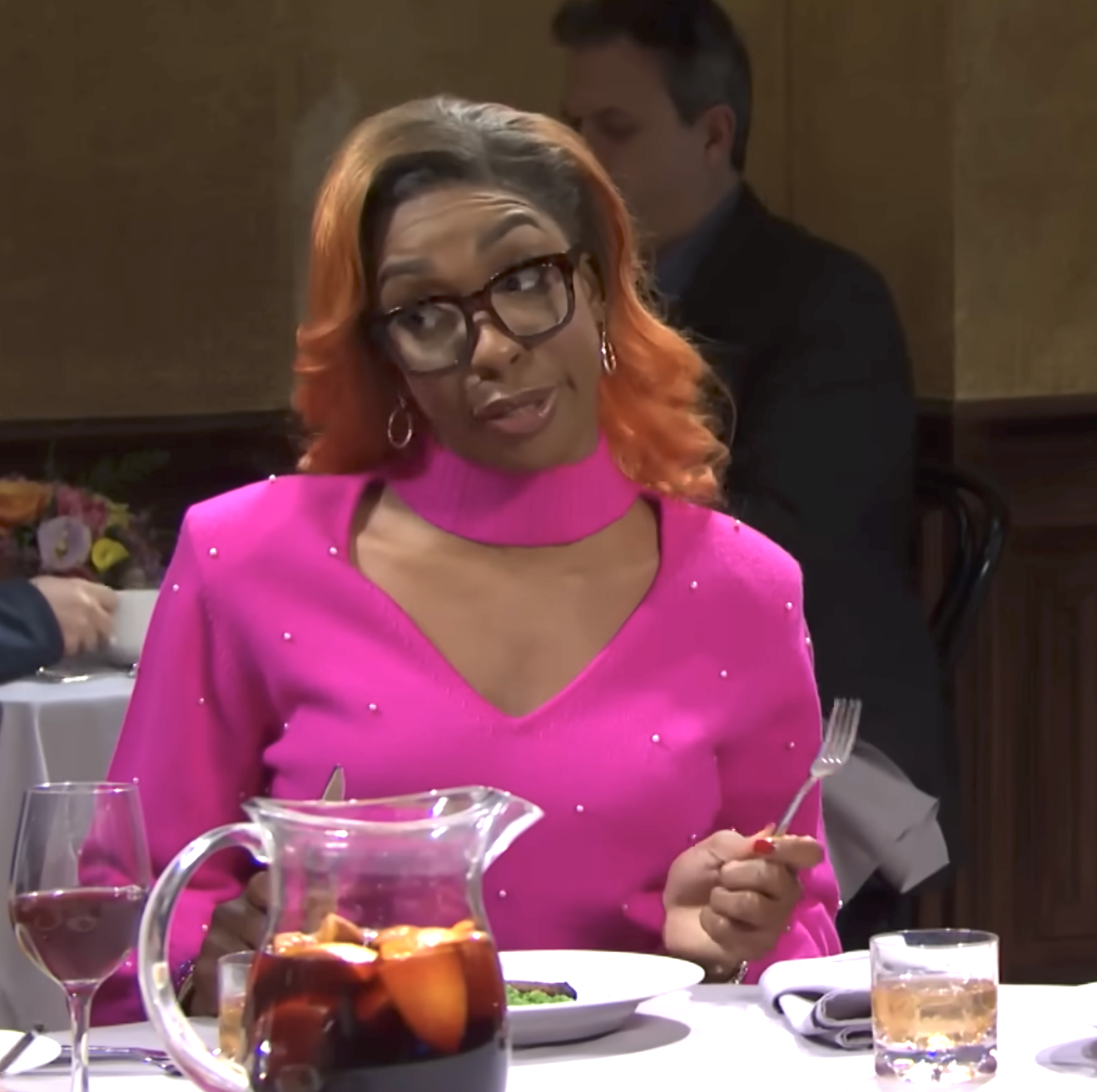 Person at a restaurant table wearing a stylish, vibrant top with glasses, holding a fork. Drinks and food on the table