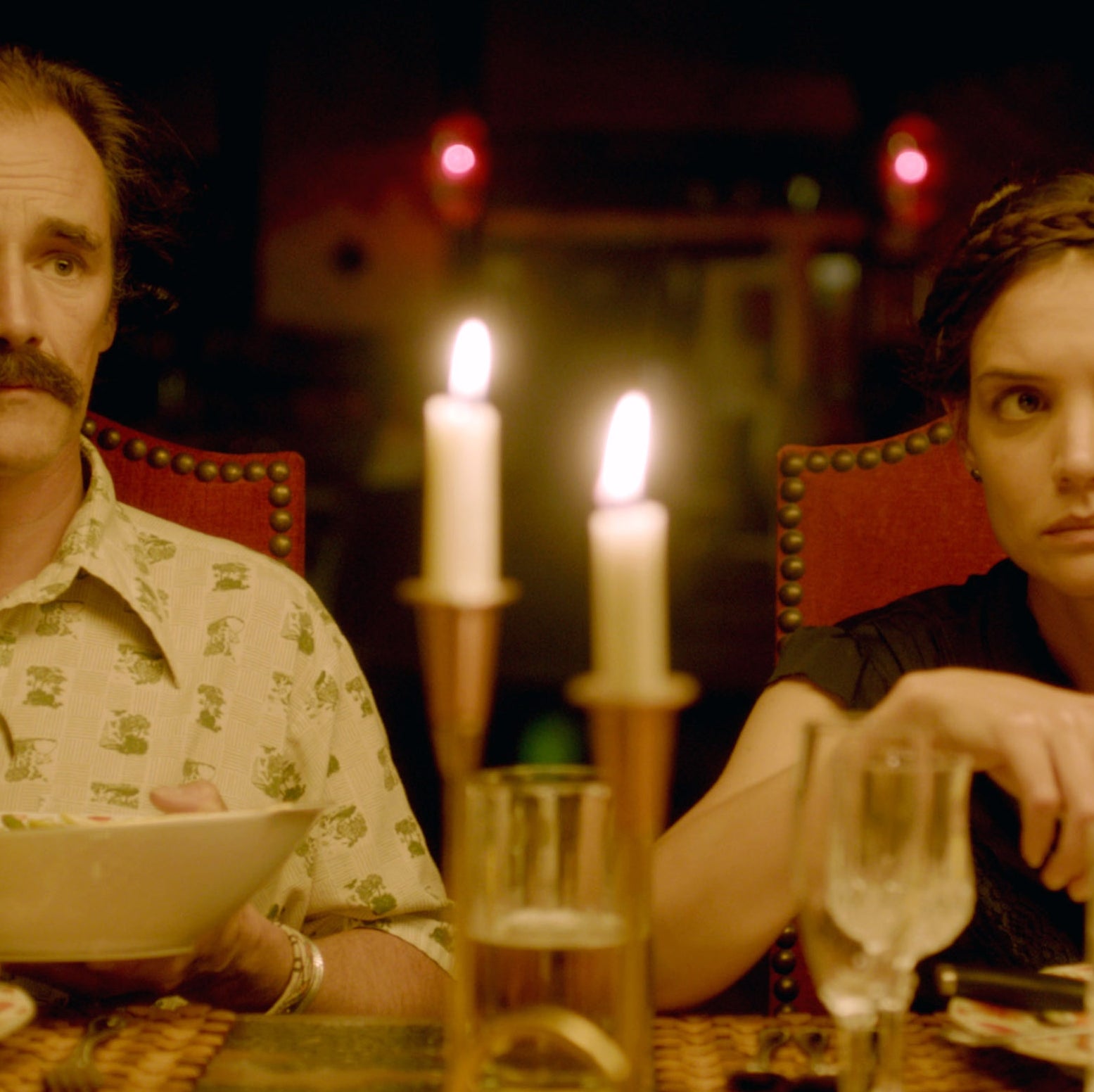Man and woman sit at a dinner table with lit candles and corn on the cob, both looking serious