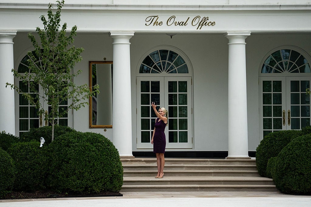 Person takes a selfie outside the entrance of a building labeled "The Oval Office," standing on steps surrounded by greenery