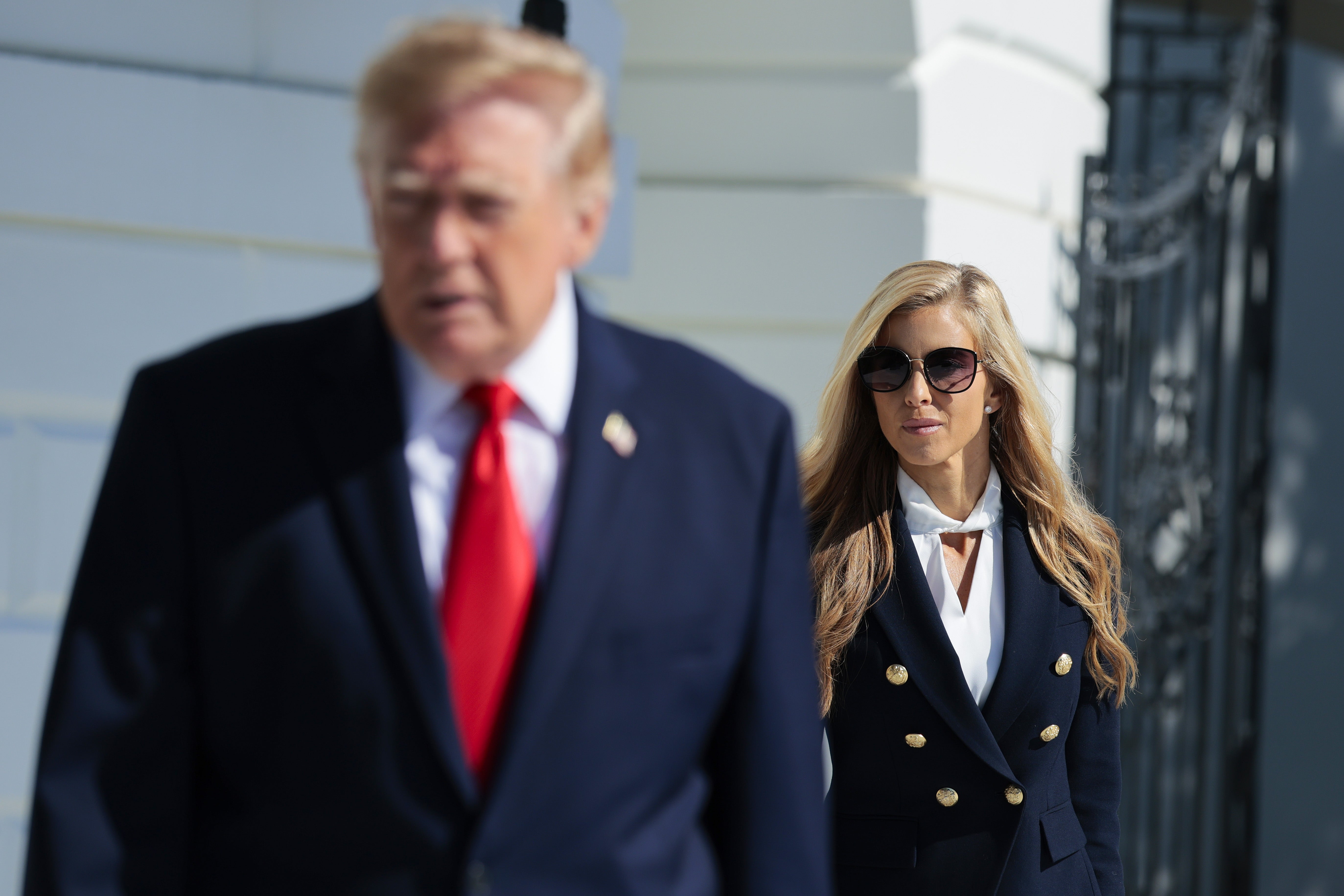 A woman in a navy double-breasted blazer and sunglasses stands on a path, while a man in a suit with a red tie walks in the foreground