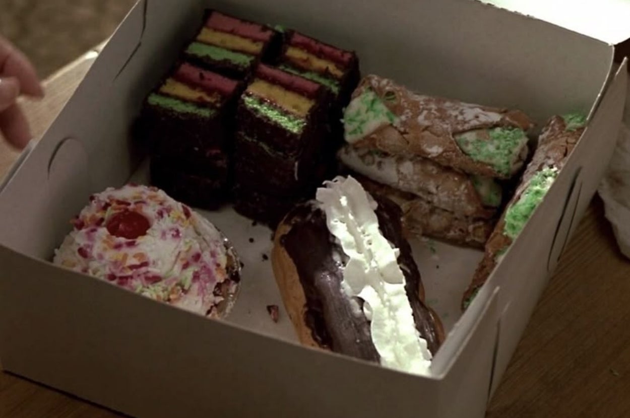 A box of assorted pastries, including a frosted eclair, cannoli, layered squares, and a cupcake with whipped cream and a cherry on top