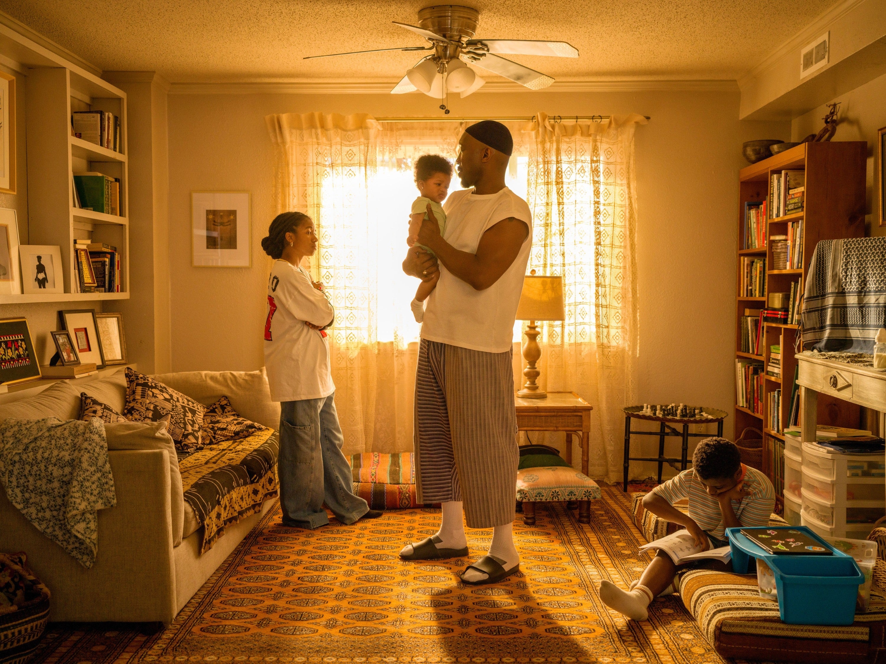 A family in a cozy living room: a person holds a baby while another stands nearby, and a child looks through a book on the floor