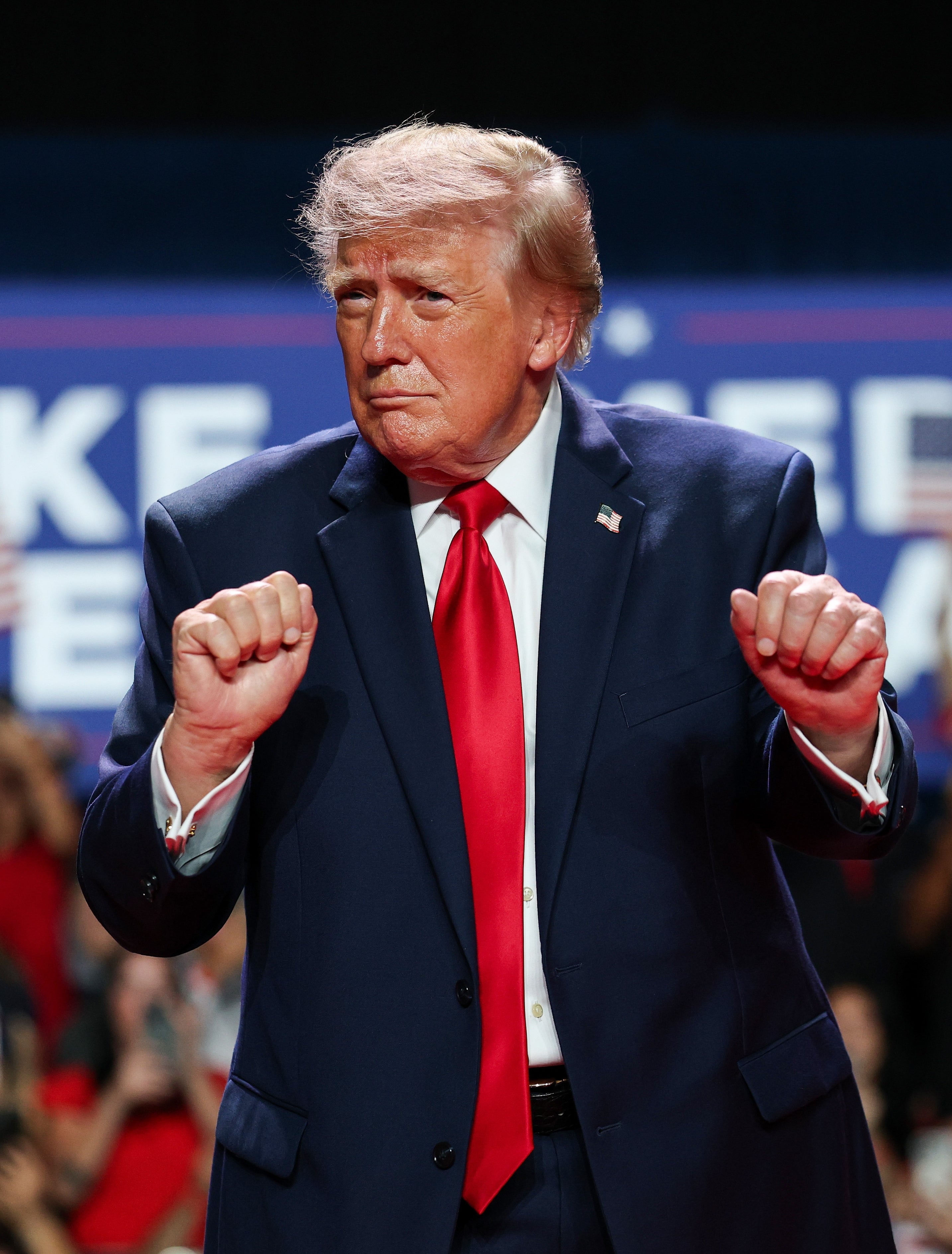 Man in suit and red tie gestures enthusiastically on stage, with a crowd and "Make America Great Again" sign in the background
