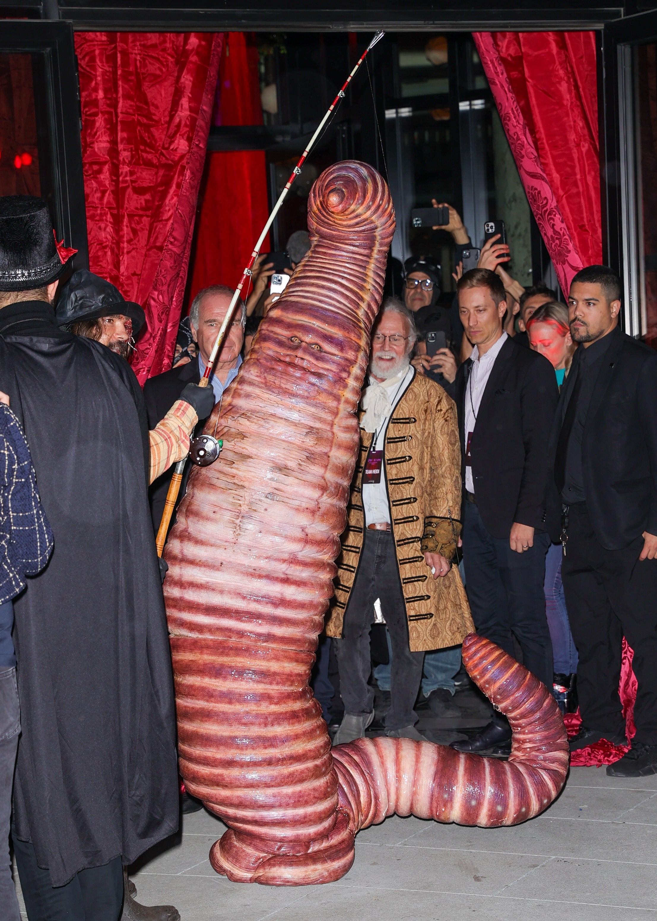 Person successful elephantine worm costume astatine an event, surrounded by onlookers, immoderate successful costumes. The mounting includes ample reddish drapes
