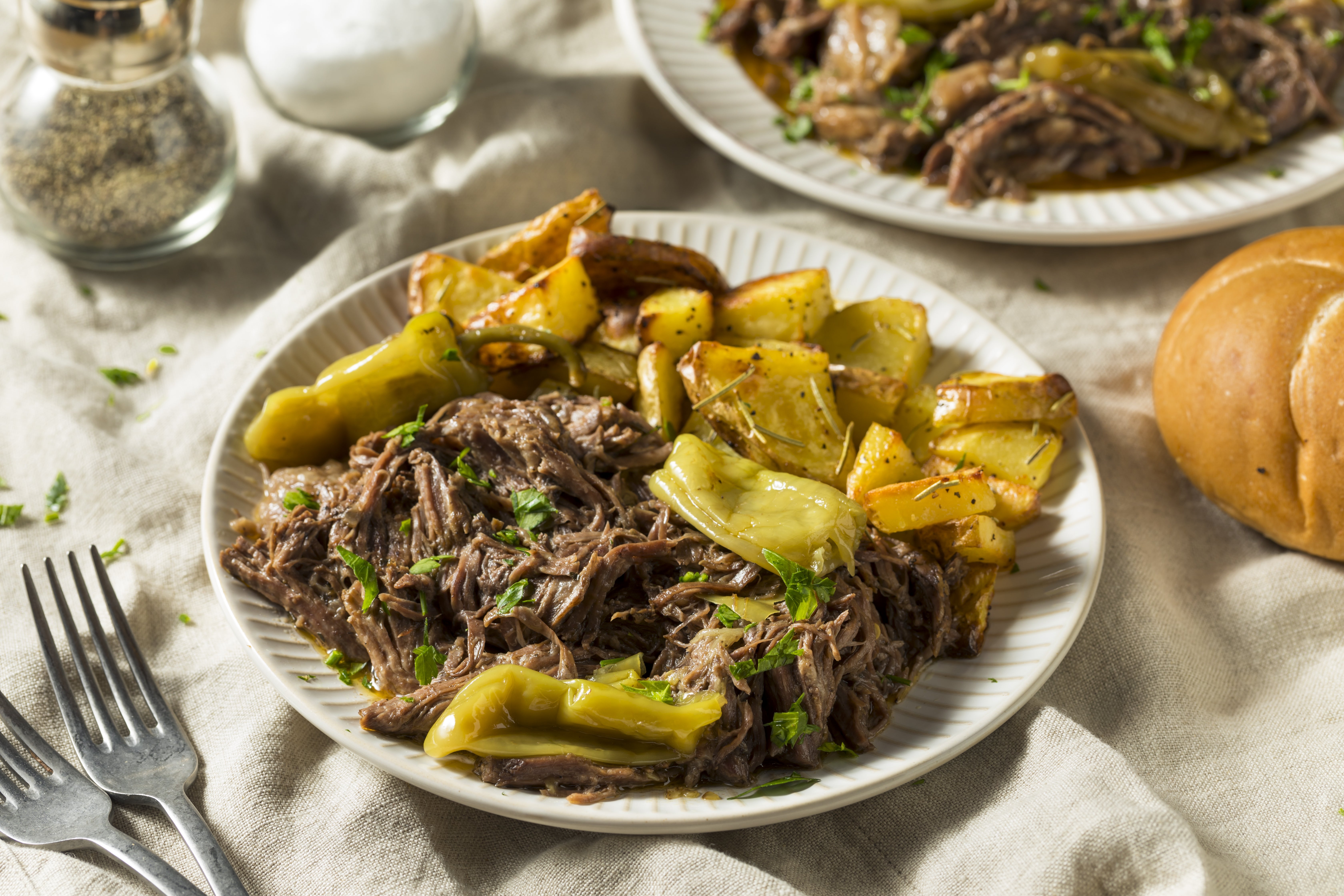 A plate of shredded beef with roasted potatoes and green peppers, garnished with herbs, next to a roll and salt and pepper shakers