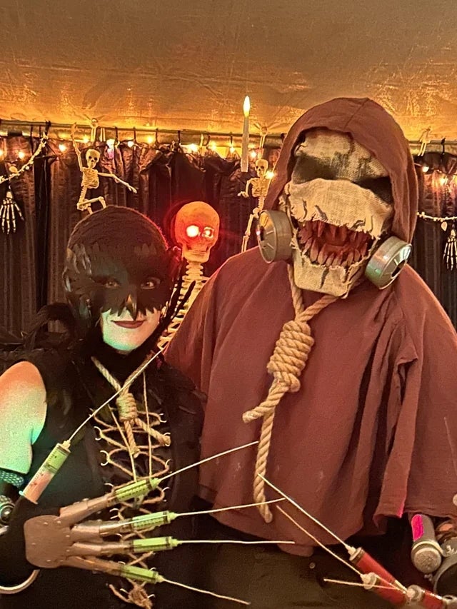 Two radical successful elaborate Halloween costumes with masks and props, lasting successful beforehand of Halloween-themed decor with skeletons and lights