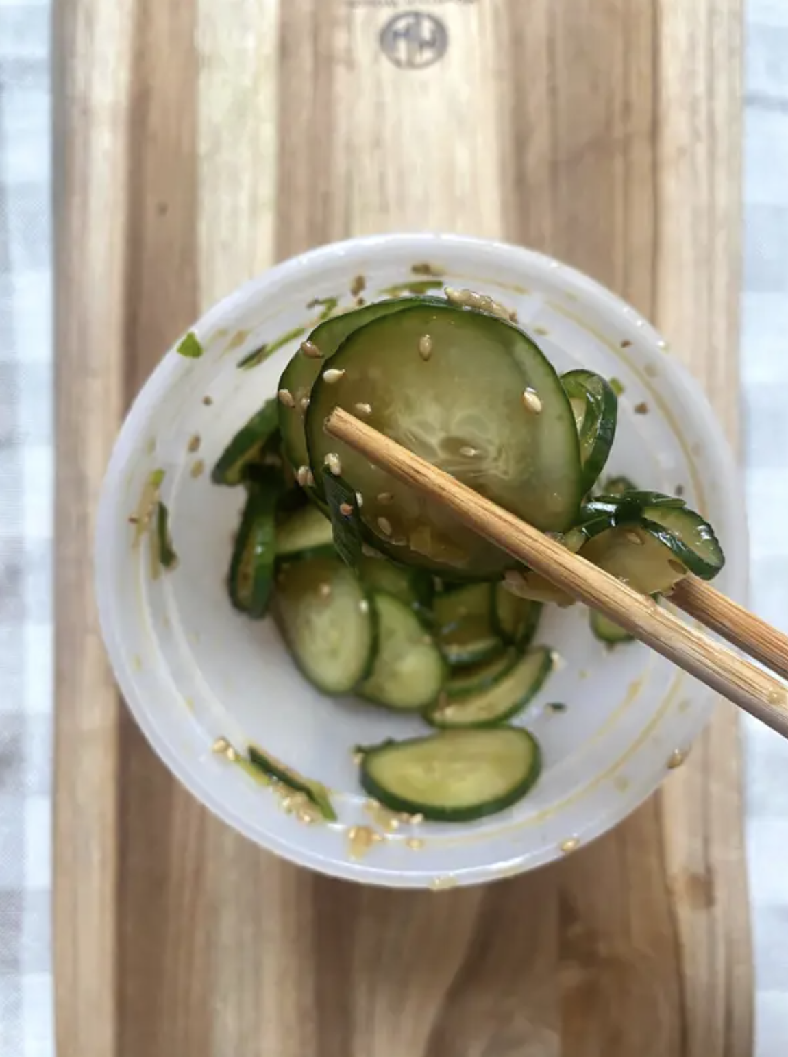 Chopsticks holding sliced cucumbers with sesame seeds over a bowl, placed on a wooden board