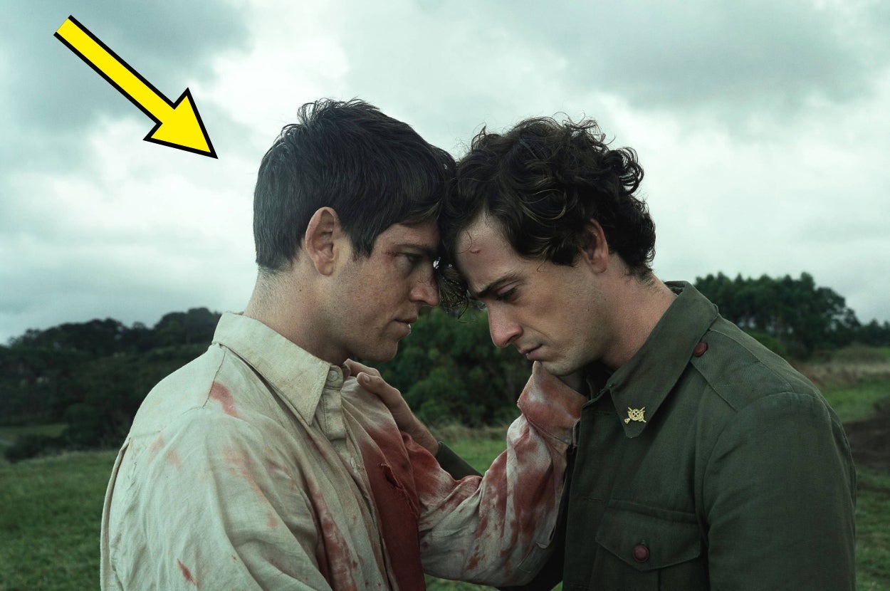 Two men with distressed expressions stand closely, one with a bloodied shirt and a patch. They have a somber, intense moment in a field