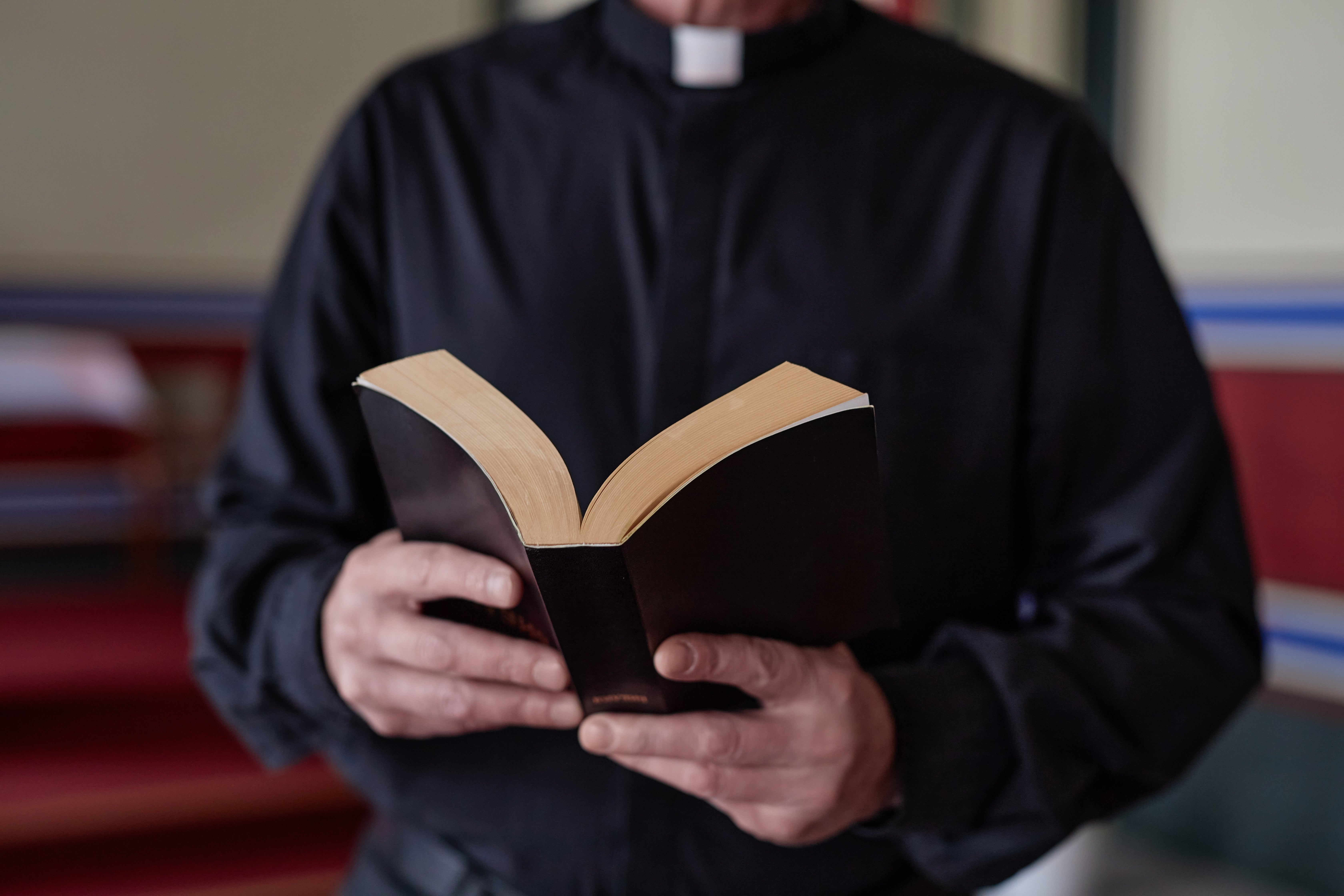 A person wearing a clerical collar holds an open book, possibly a Bible, while standing indoors
