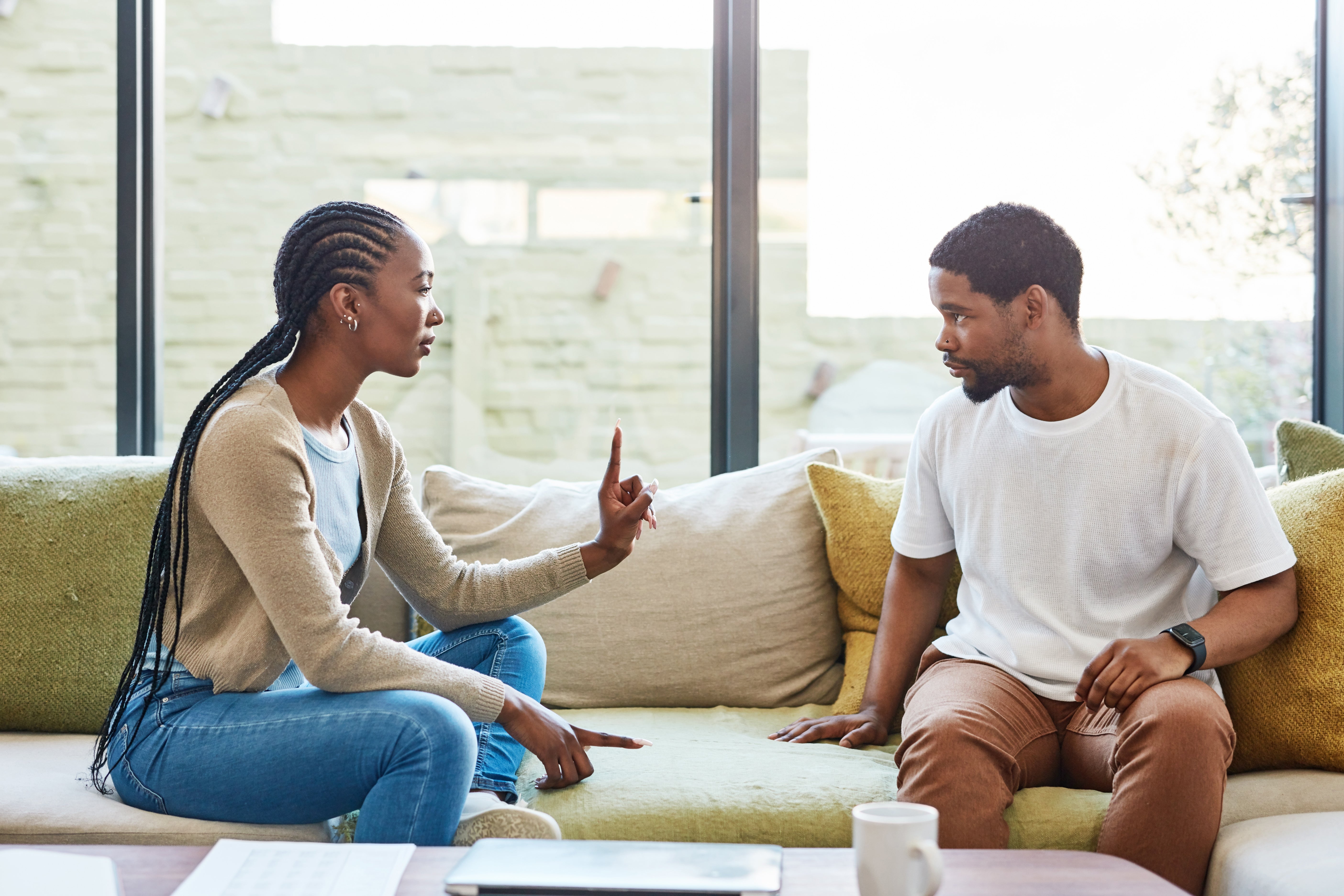 Two people sit on a couch having a serious conversation. One is gesturing with a finger, while the other listens intently