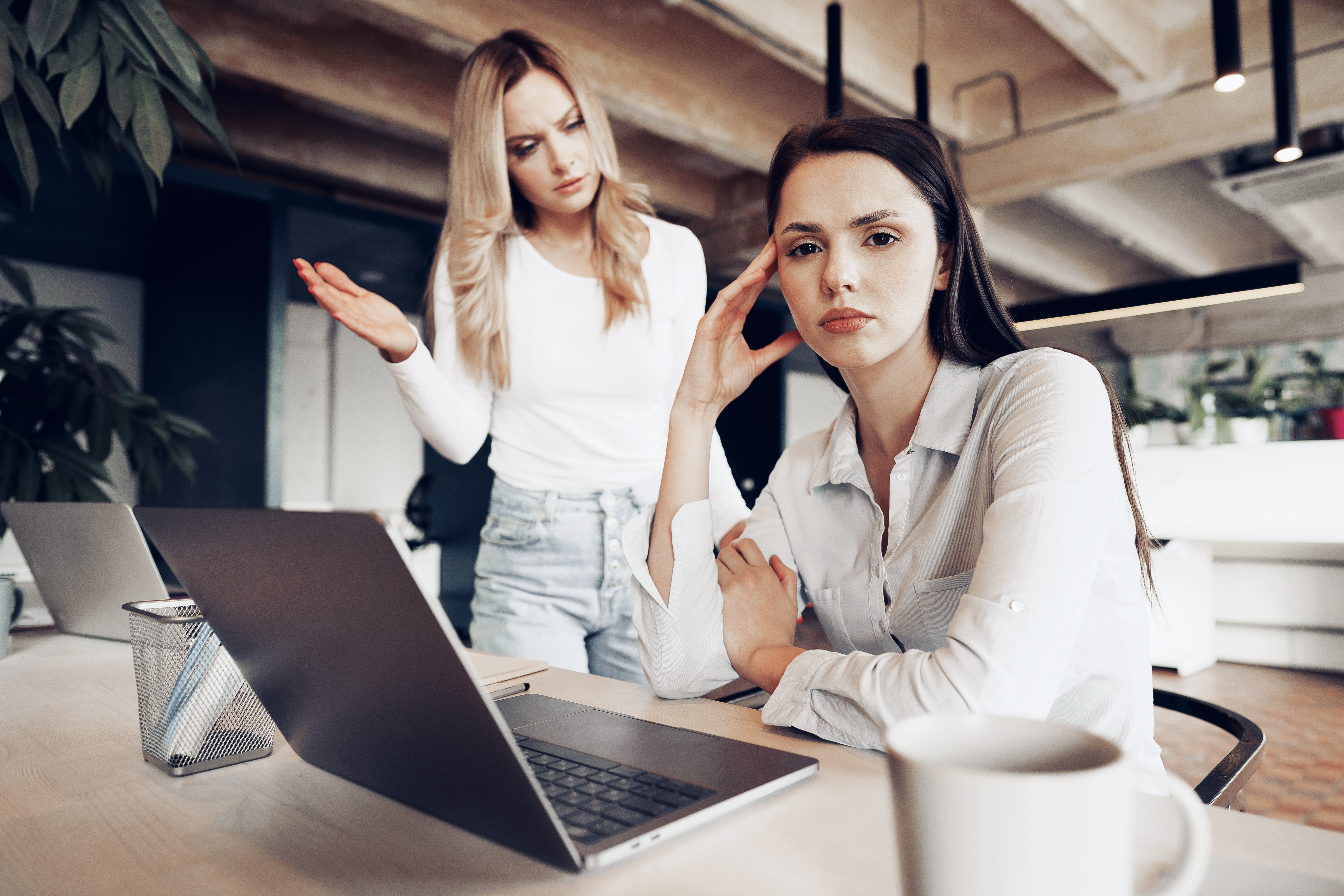 Two women are in an office setting with laptops. One is sitting, looking frustrated, while the other stands, gesturing as if explaining something