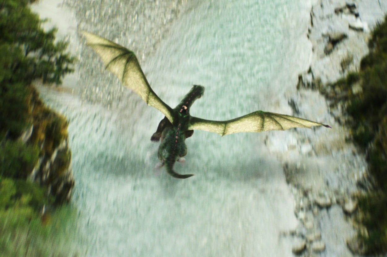 A dragon from "Pete's Dragon" with large, outstretched wings flies over a rocky, river-filled landscape