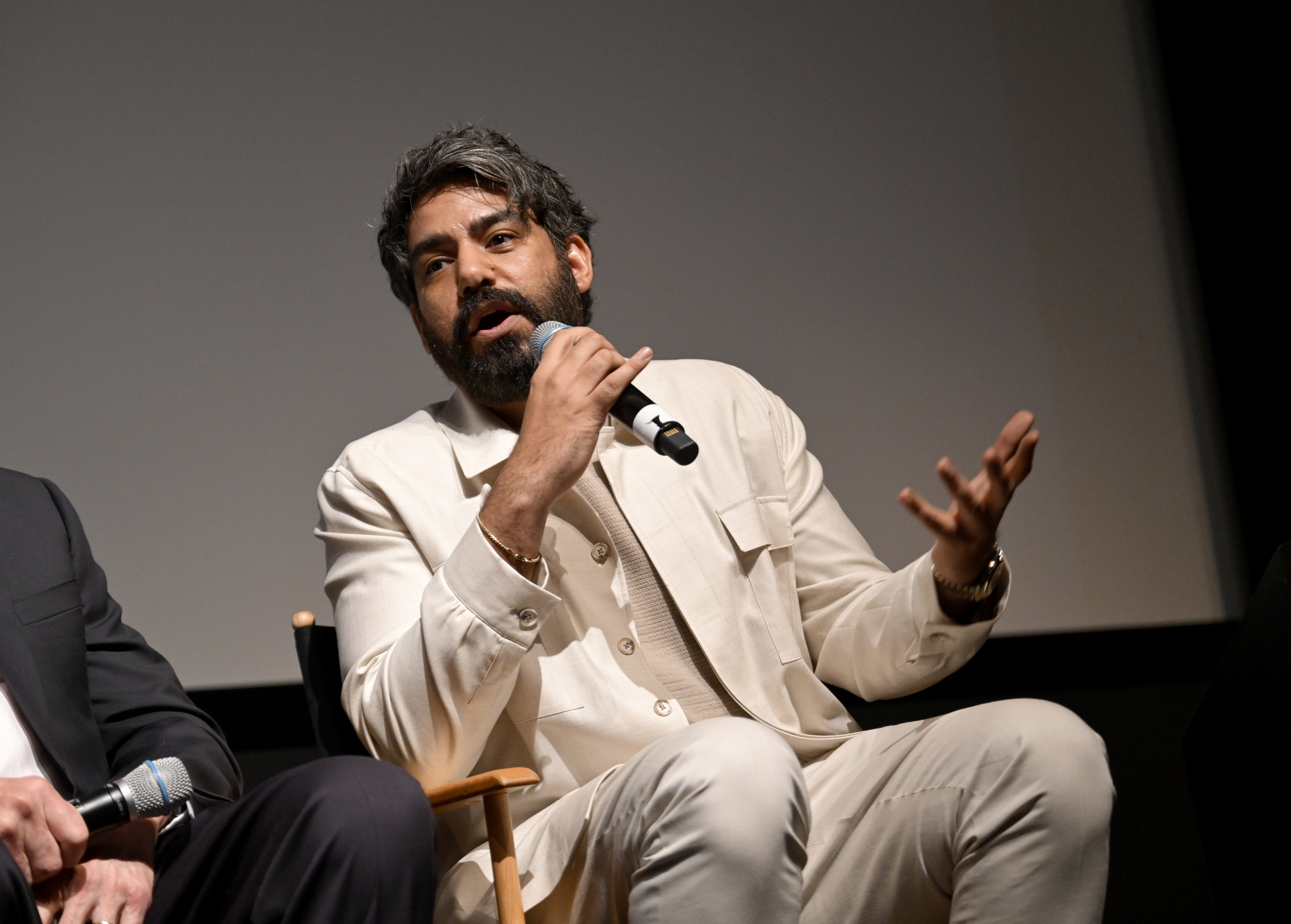 Rahul Kohli sitting and speaking in a panel discussion, wearing a light suit, holding a microphone, gesturing with one hand