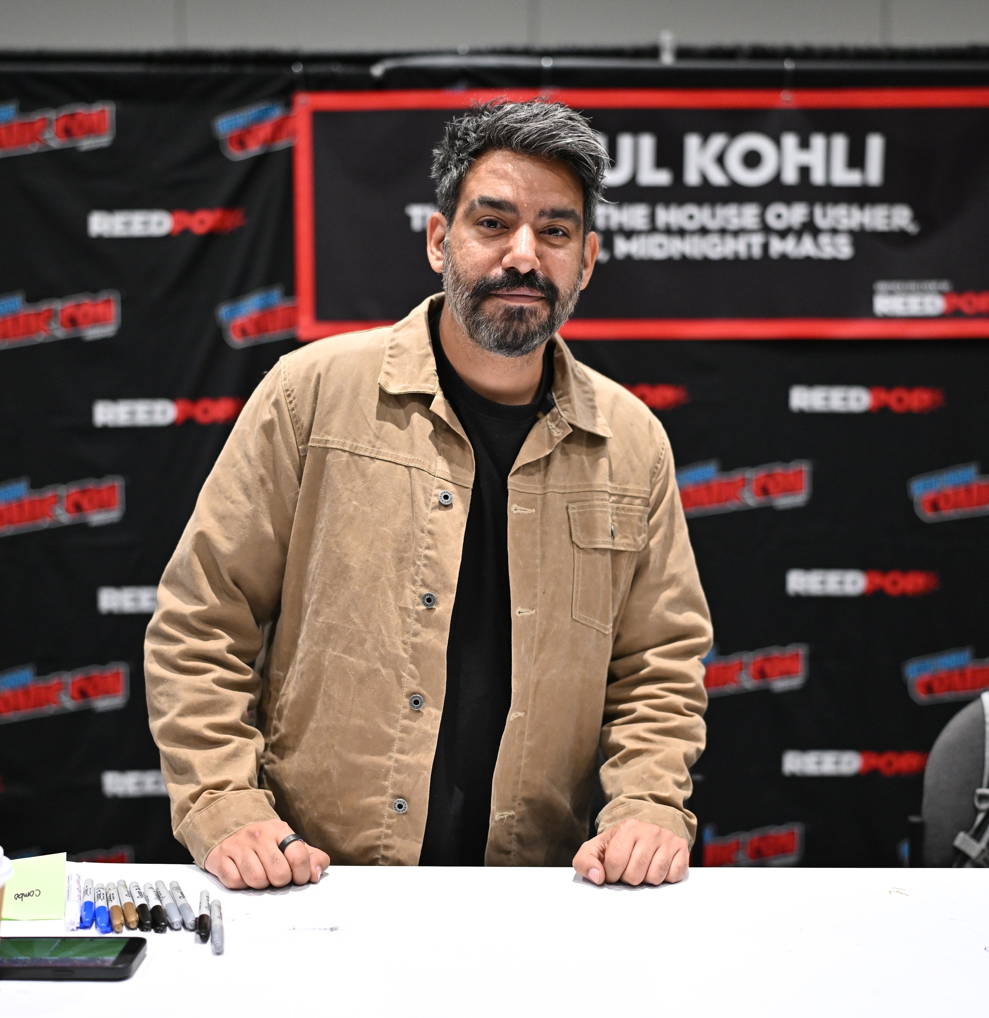 Rahul Kohli at a NYCC convention booth, wearing a casual jacket, standing behind a table with markers and a coffee cup. Background banners display the convention's logos.