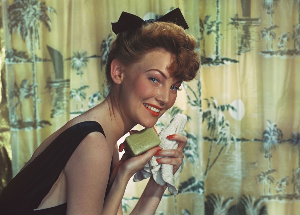 Woman smiling while holding soap and towel, styled in vintage fashion with pinned-up hair and a sleeveless dress, against a patterned curtain backdrop