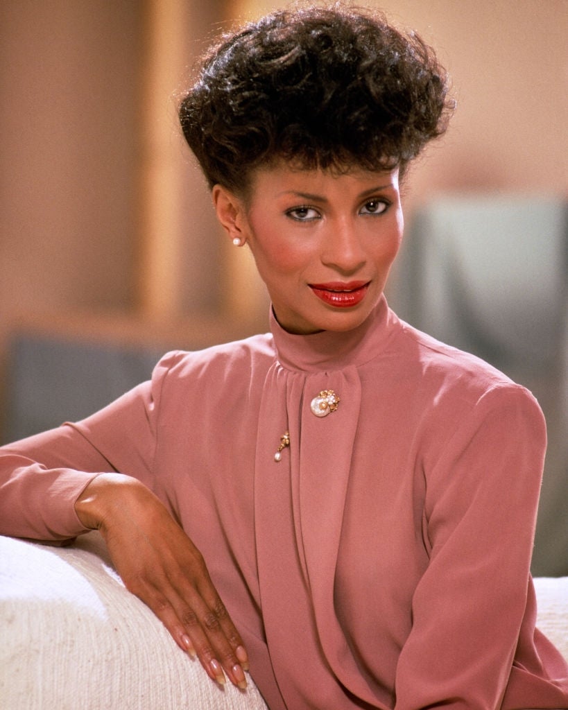 Person with styled short hair wearing a high-neck blouse with a brooch, seated indoors, looking confidently at the camera