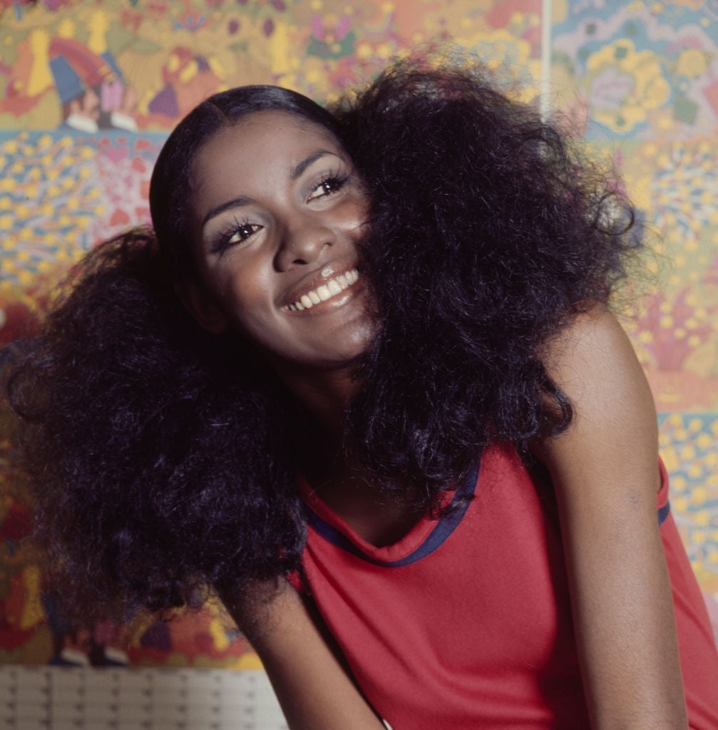 Woman smiling with voluminous hair, wearing sleeveless top in front of vibrant patterned backdrop. Retro, joyful expression