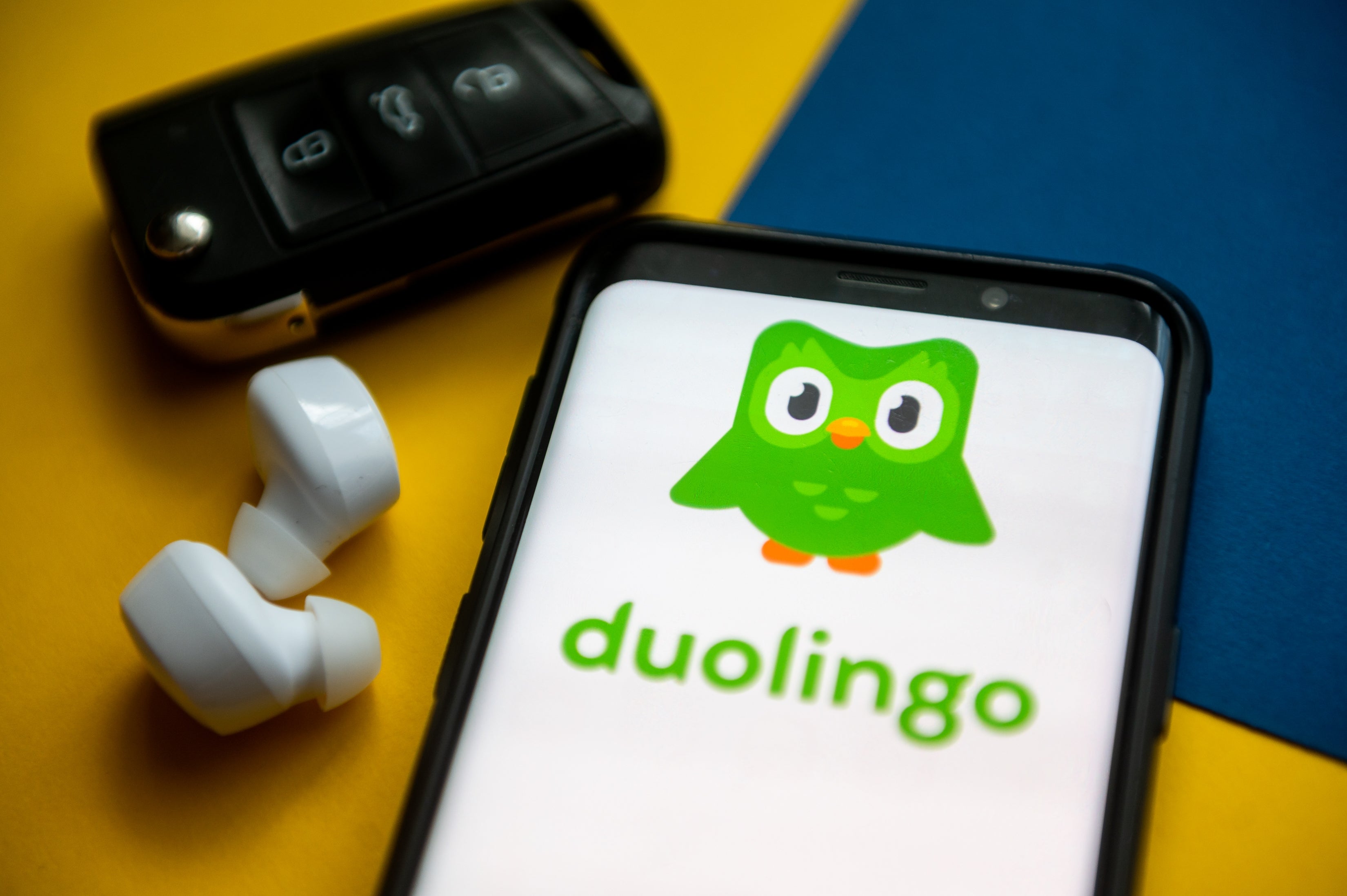 Phone showing Duolingo app logo with owl mascot adjacent to earbuds and a car key