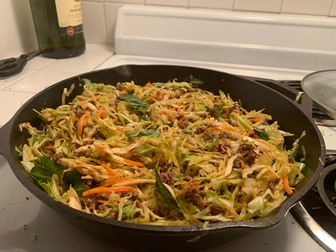 A skillet filled with a cooked dish of shredded cabbage, carrots, ground meat, and greens on a stovetop