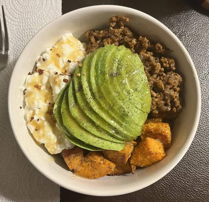 A bowl with ground meat, sliced avocado, roasted sweet potatoes, and cottage cheese sprinkled with seasoning