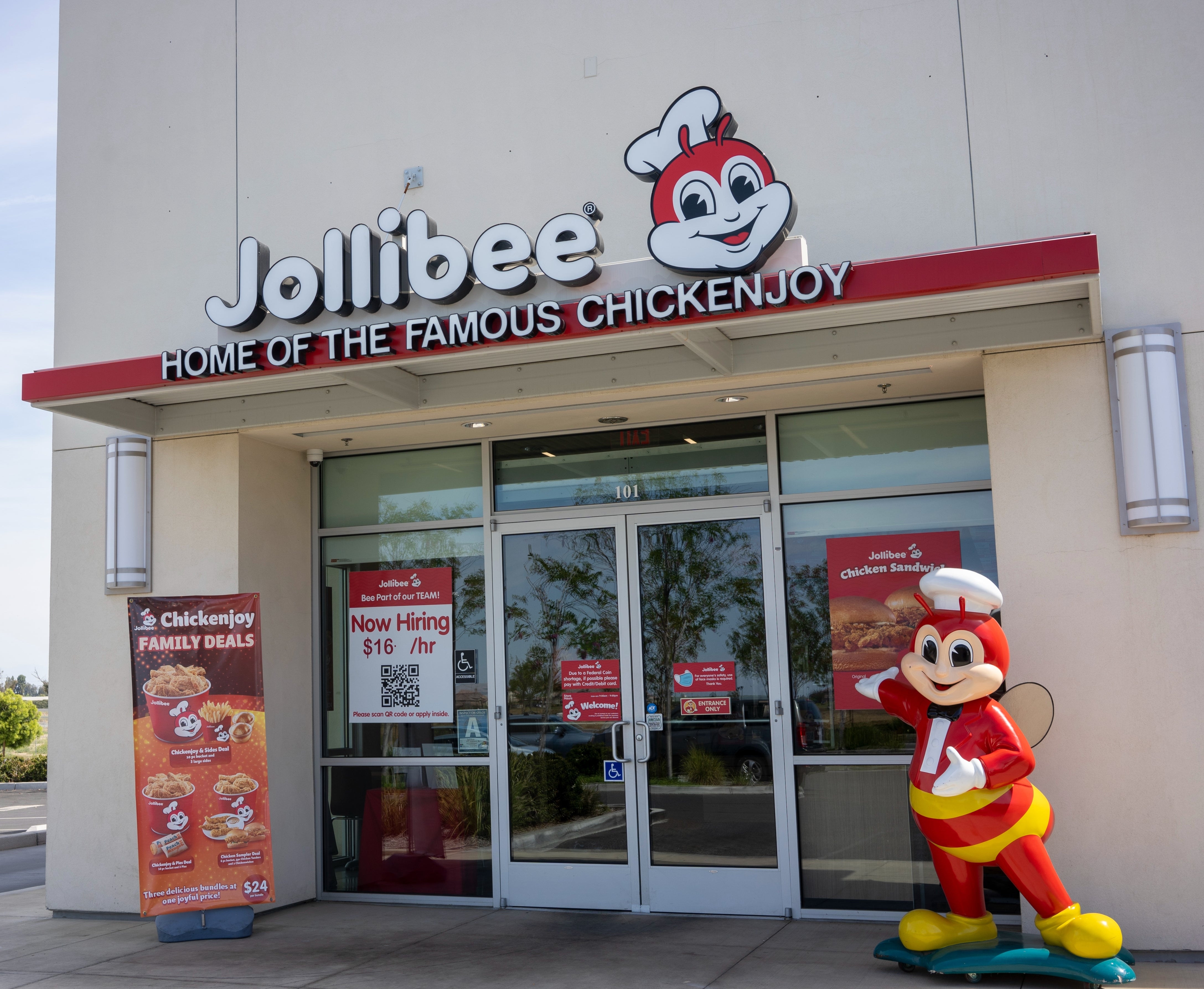 Jollibee fast-food edifice entranceway with a mascot statue outside, signage advertizing "Now Hiring" astatine $16/hr, and household repast deals