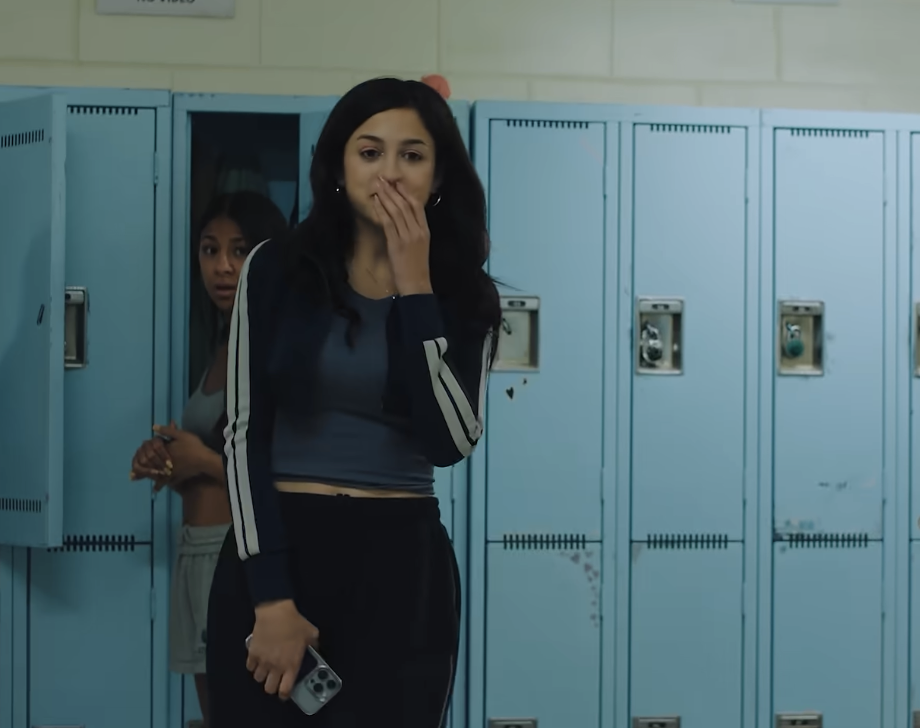 Josie Totah stands in front of open lockers, hand covering her mouth, holding a smartphone