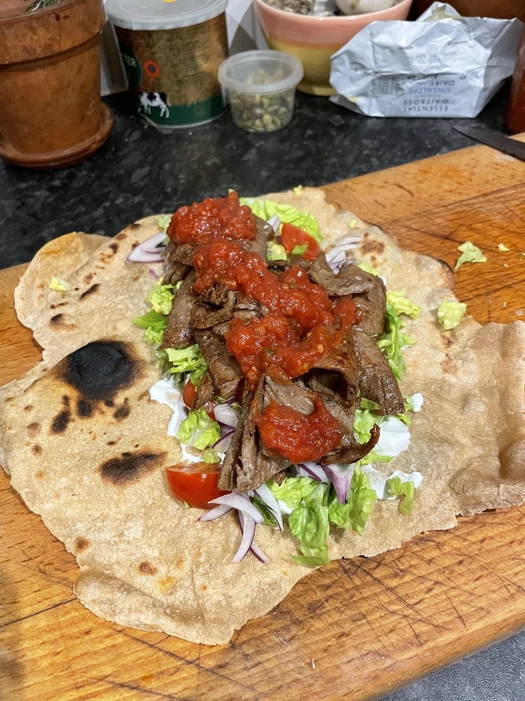 Flatbread topped with sliced meat, lettuce, tomatoes, onions, and a dollop of sauce on a wooden cutting board