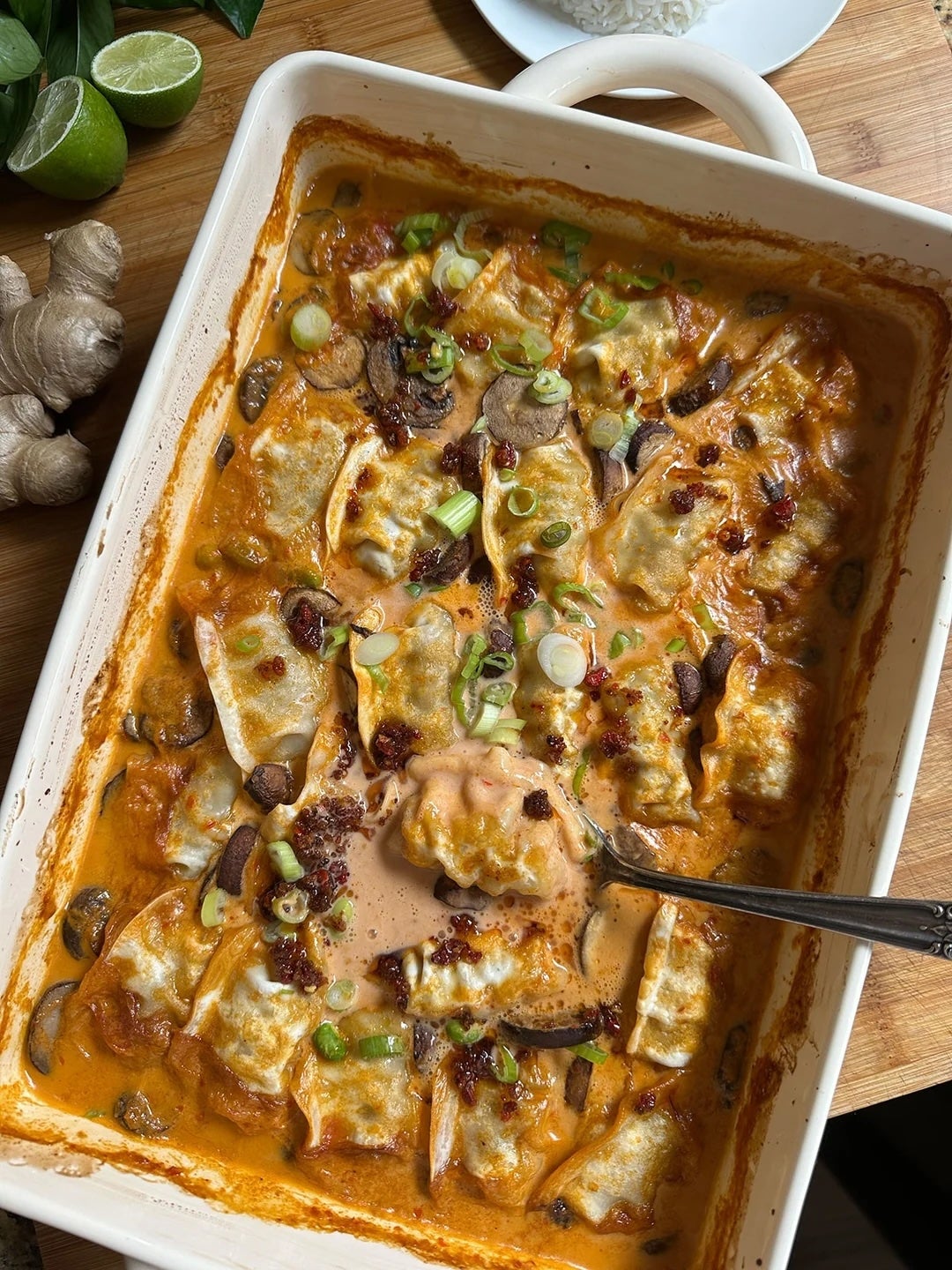 Baked dumplings in a dish with creamy sauce, topped with scallions and bacon bits. Ginger and lime visible nearby
