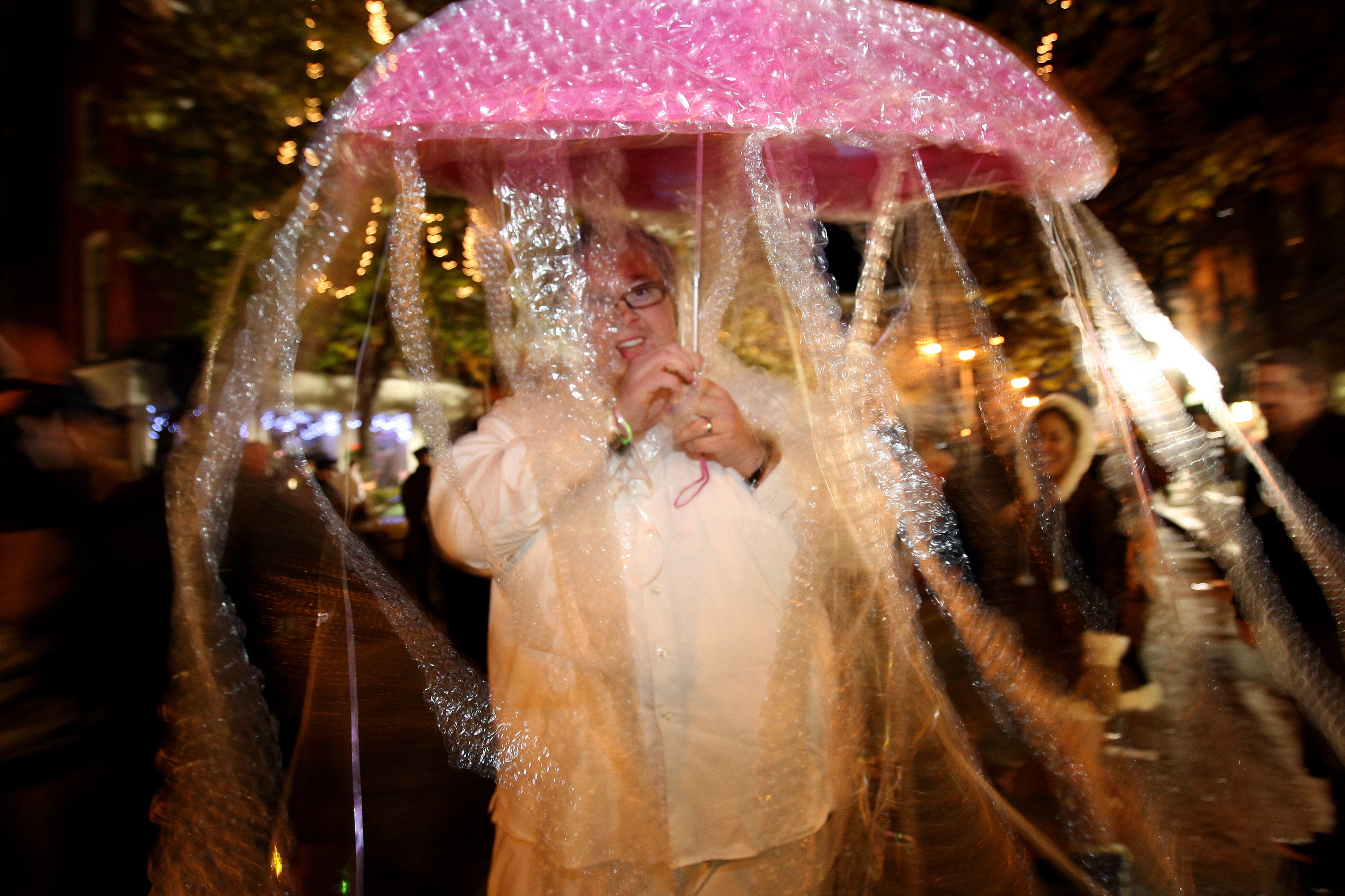 Person dressed successful a originative jellyfish costume utilizing bubble wrapper and a pinkish umbrella, walking connected a engaged thoroughfare during a nighttime event