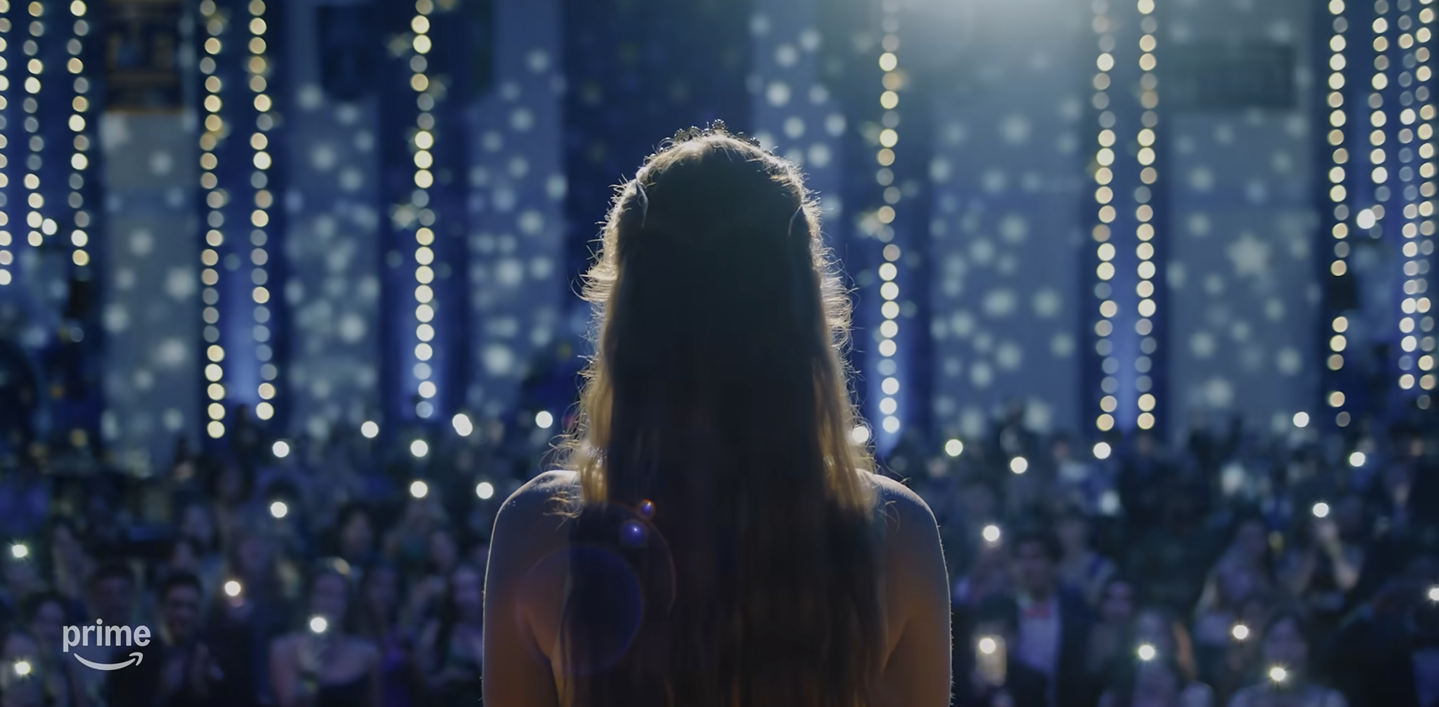 Carrie stands on stage with sparkling lights in the background, facing an audience that's illuminated by phone lights