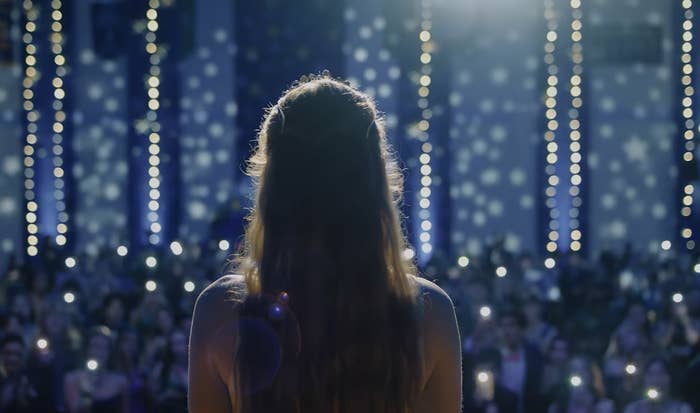 Carrie stands on stage with sparkling lights in the background, facing an audience that's illuminated by phone lights