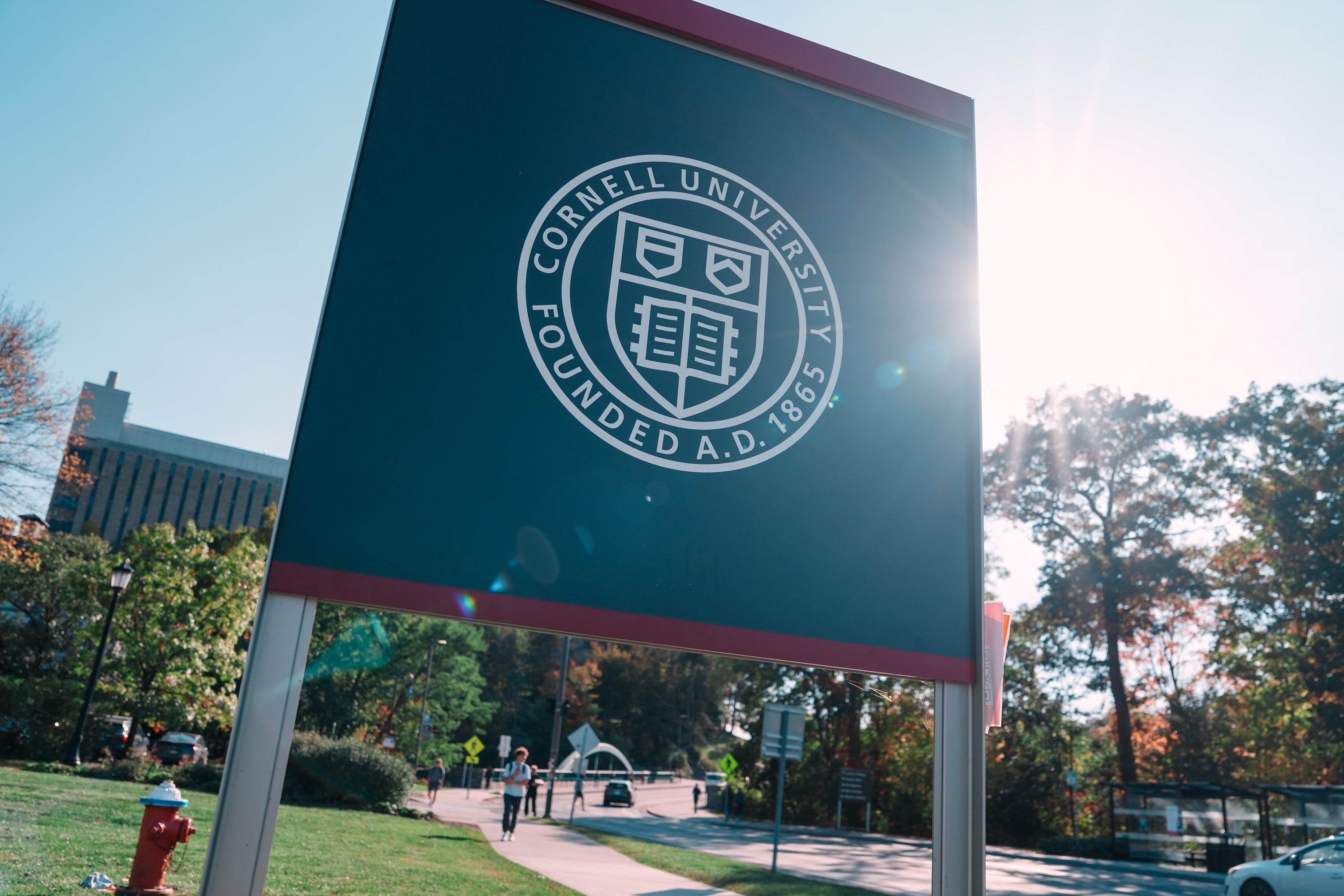 Sign at Cornell University campus entrance with the university emblem, surrounded by autumn trees and a distant clear sky