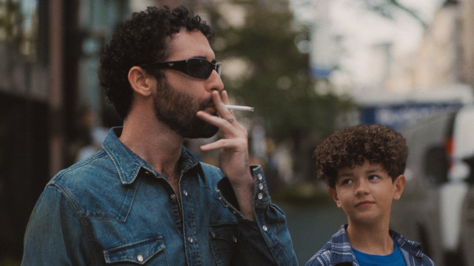 Man in denim shirt smoking and wearing sunglasses, stands beside a child in a plaid shirt, looking up at him. Urban background