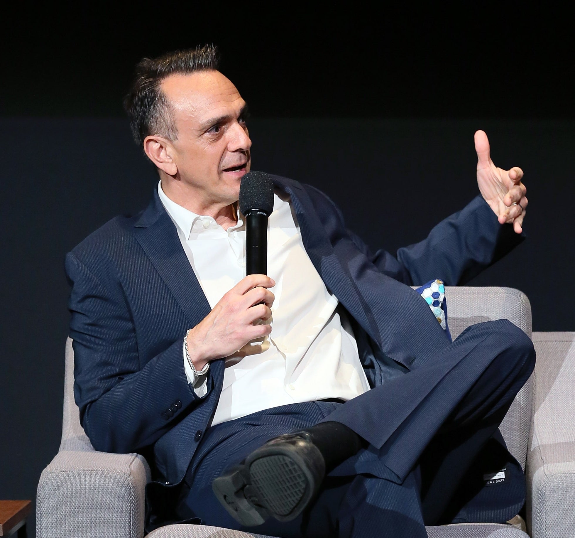 Actor Hank Azaria successful a suit sits connected a couch, holding a microphone and gesturing with 1 manus during an interview.