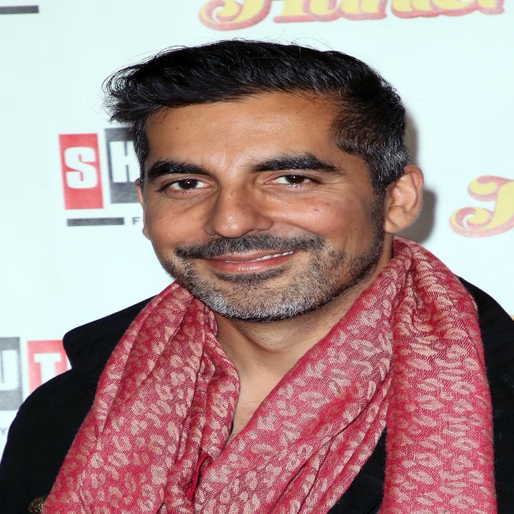 Sunil Malhotra wearing a black jacket and patterned scarf smiles at a media event backdrop with logos