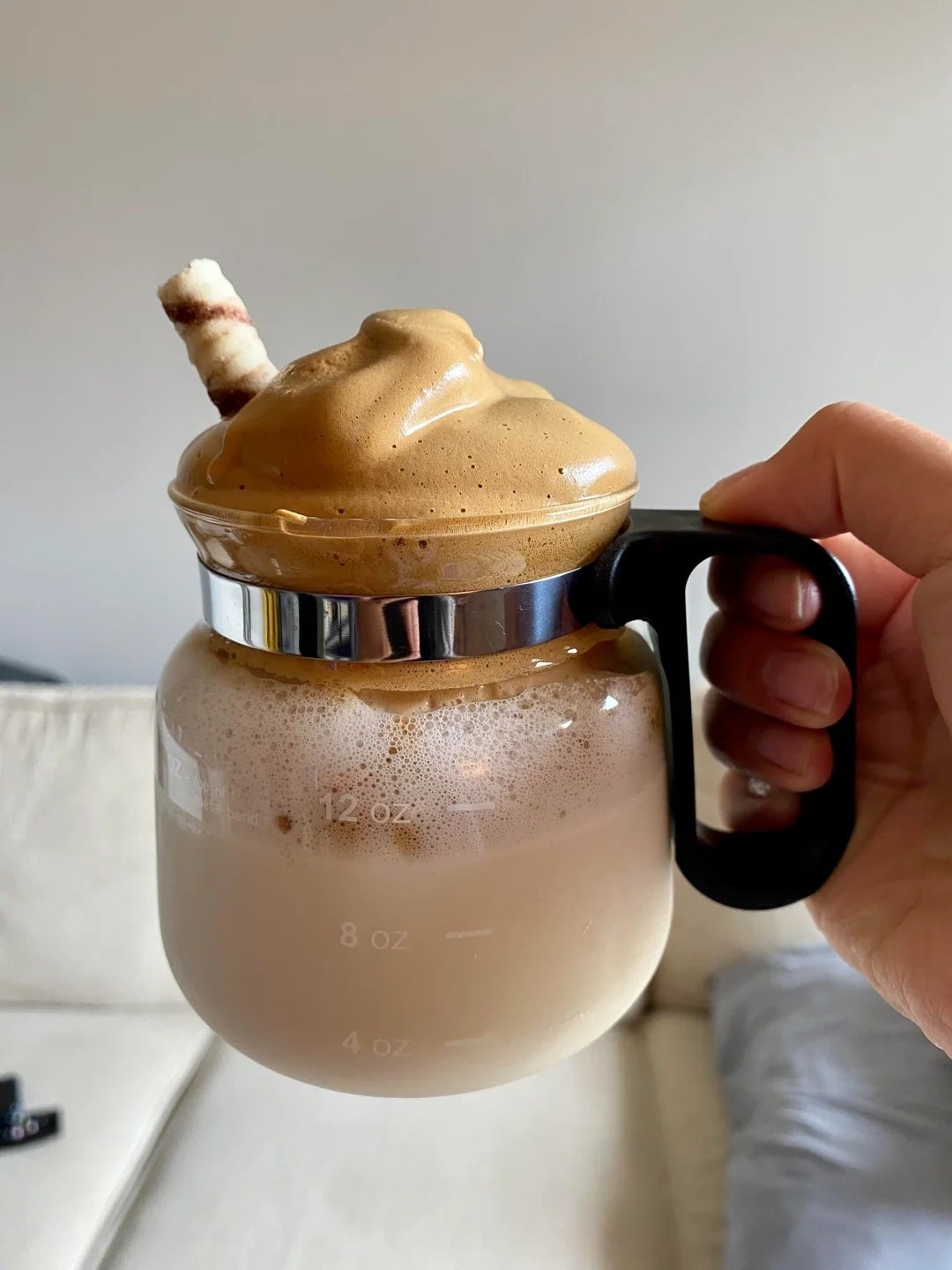 A hand holds a glass jug with creamy whipped coffee on top, garnished with a wafer stick