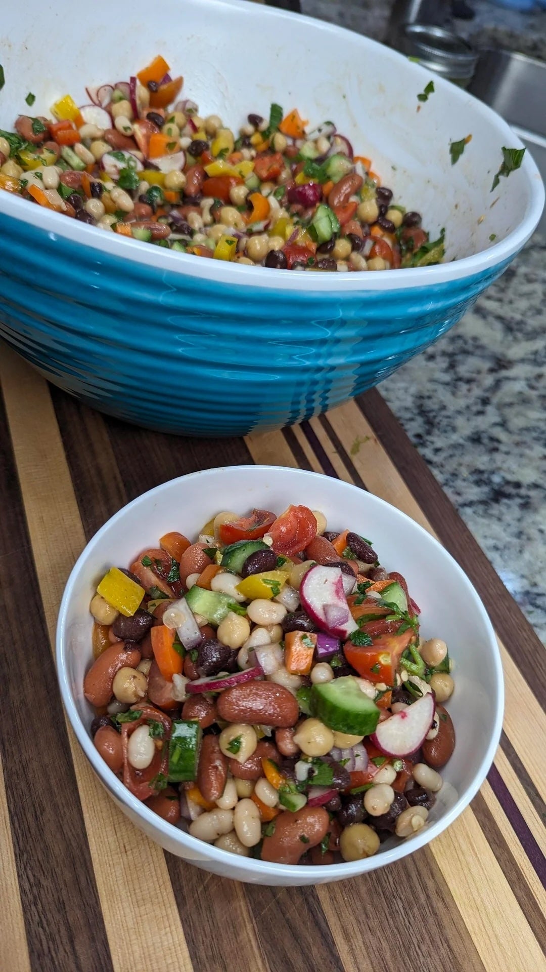 A bowl filled with a colorful mixed bean salad with vegetables on a wooden countertop, beside a larger serving bowl