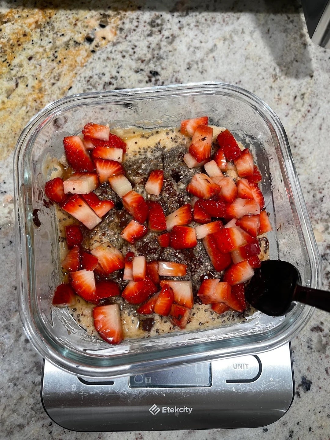 A glass container on a scale holds a mixture topped with chopped strawberries and chia seeds, likely preparing a dessert or breakfast dish