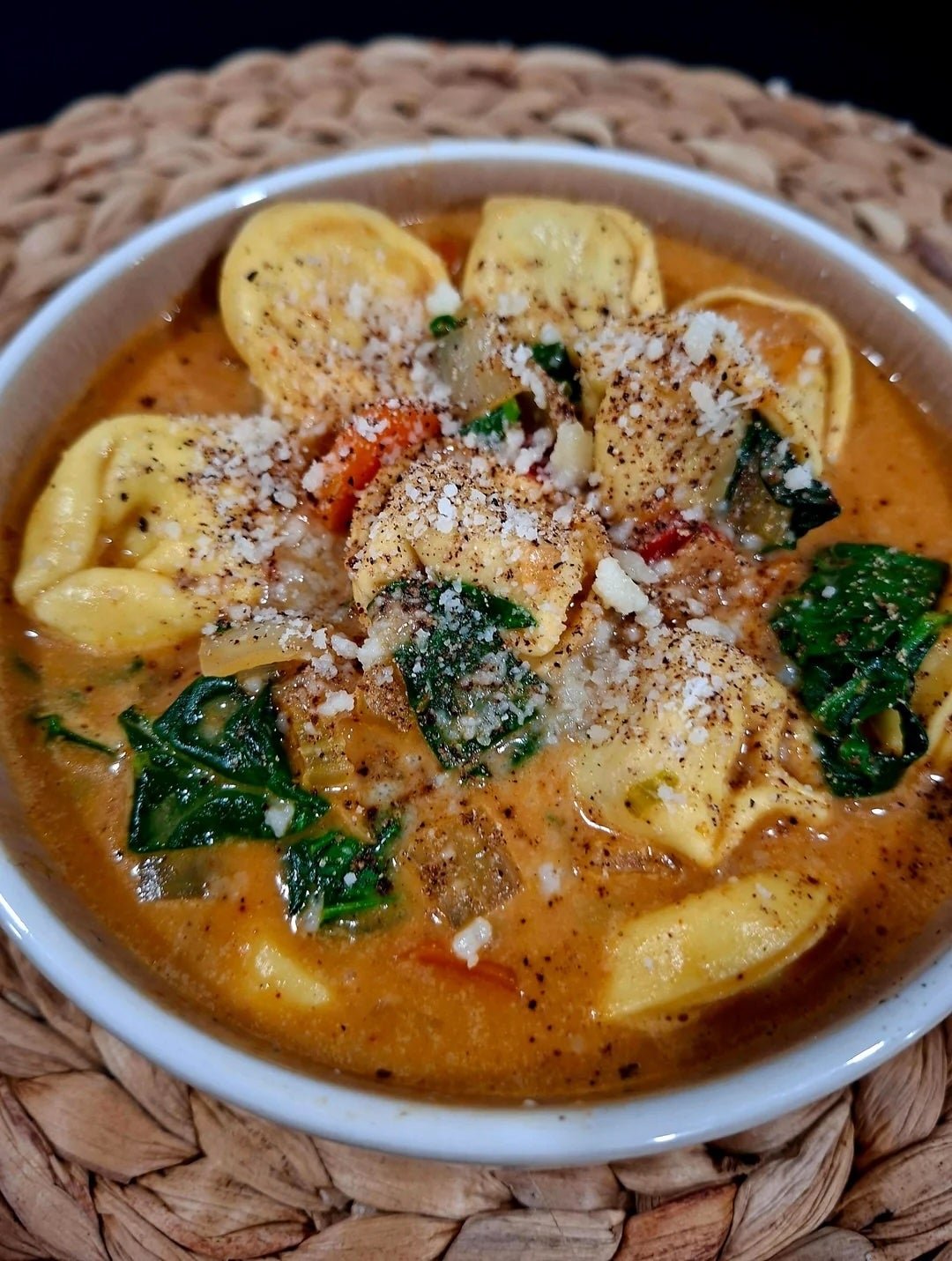 A bowl of creamy tortellini soup with spinach, tomatoes, and sprinkled cheese on a woven placemat