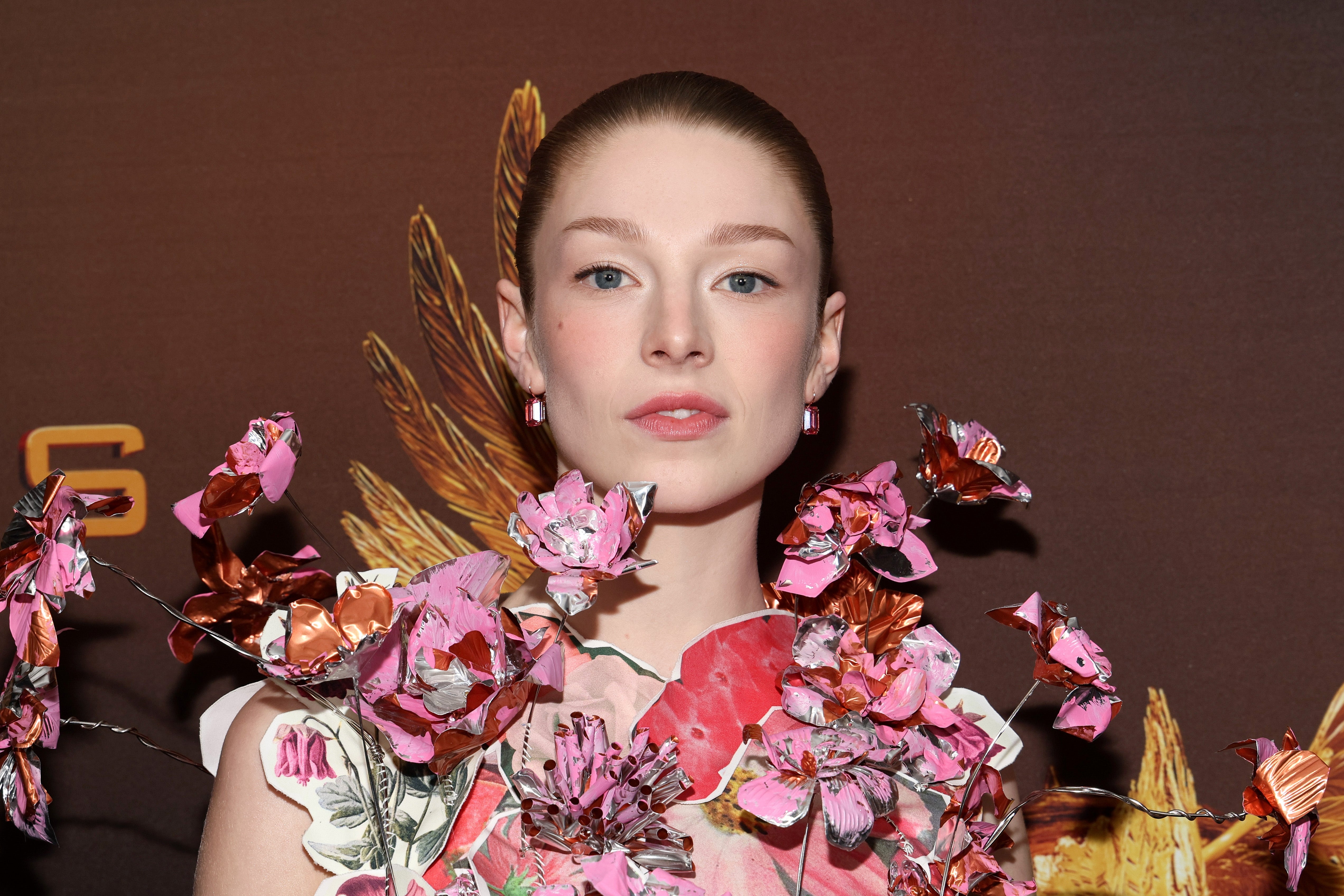 Person on red carpet wearing floral-themed outfit with intricate flower details. Hair is tied back, and they are looking directly at the camera