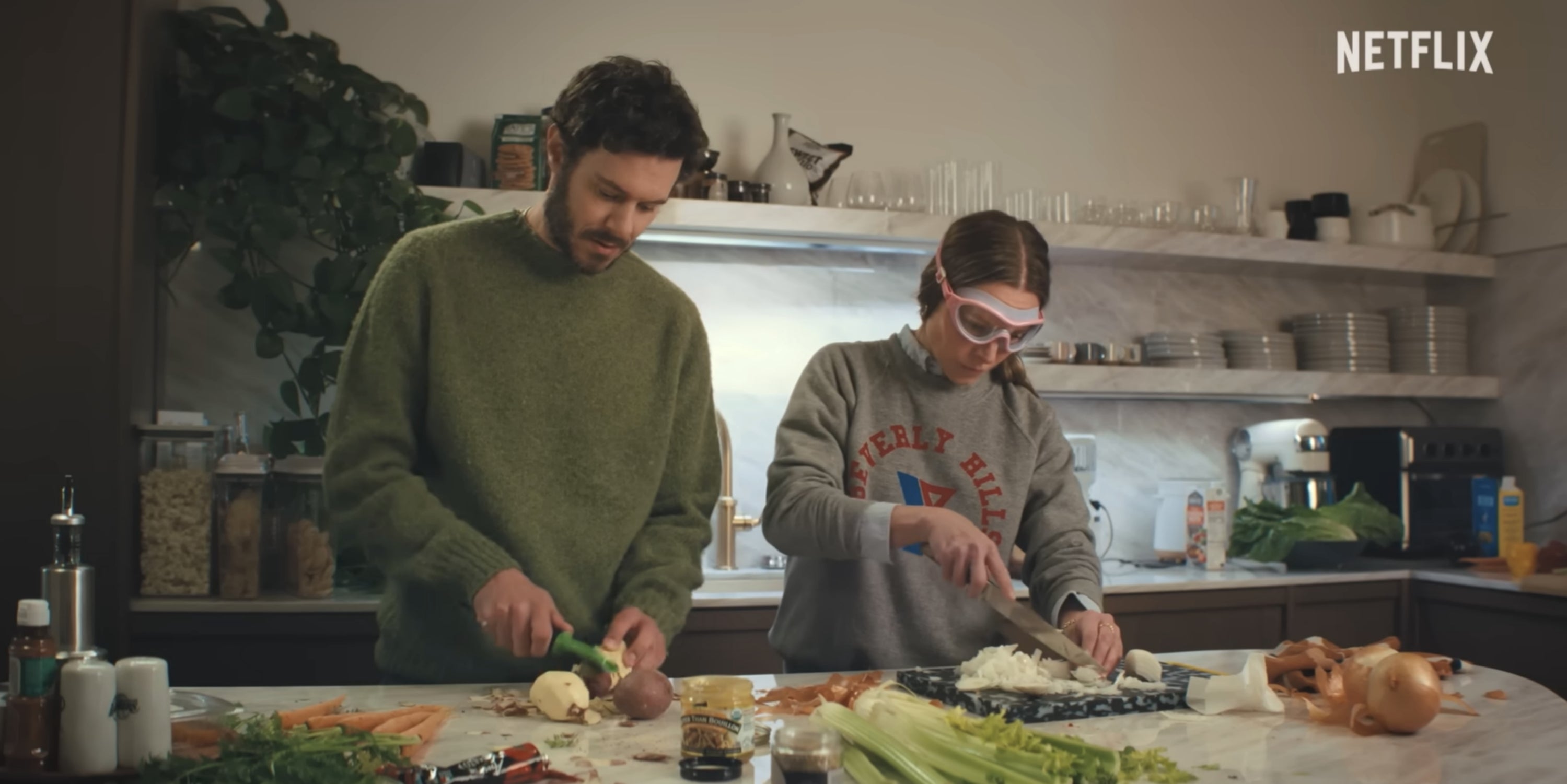 Two people cooking in a kitchen, chopping vegetables. The woman wears protective goggles while slicing onions. Shelves with dishes in the background